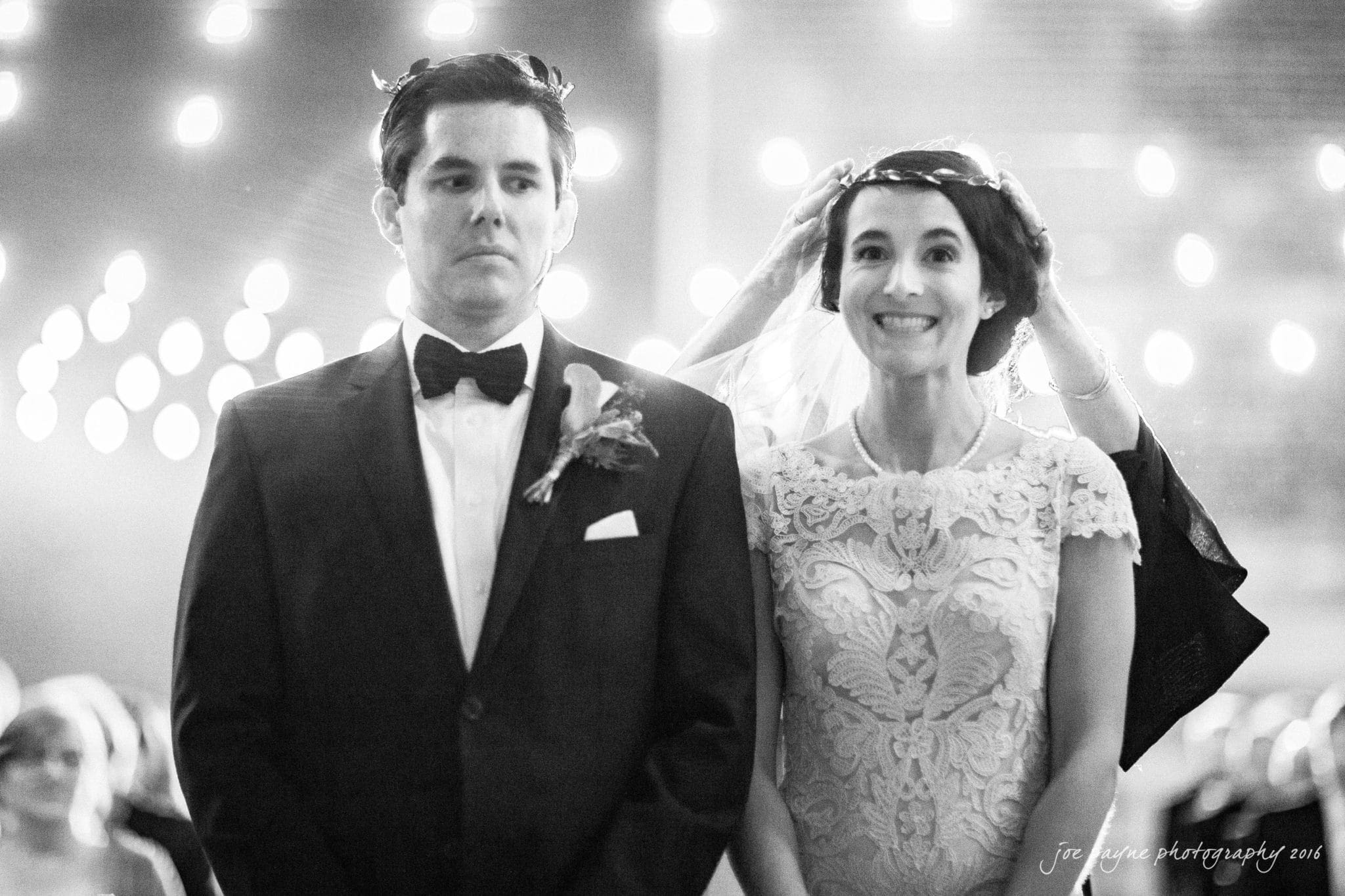 cloth mill at eno river wedding - eleanna & michael A black and white photo of a wedding ceremony. A bride in a lace dress and a groom in a tuxedo stand side by side. The bride smiles brightly while the groom has a serious expression. Soft, twinkling lights illuminate the background.