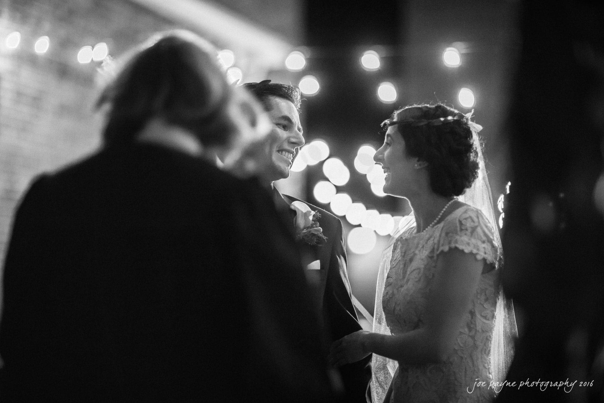 cloth mill at eno river wedding - eleanna & michael A black-and-white photo shows a couple smiling at each other during their wedding ceremony. They stand under string lights, with a blurred officiant in the foreground. The bride is wearing a lace gown, and the groom is in a suit.