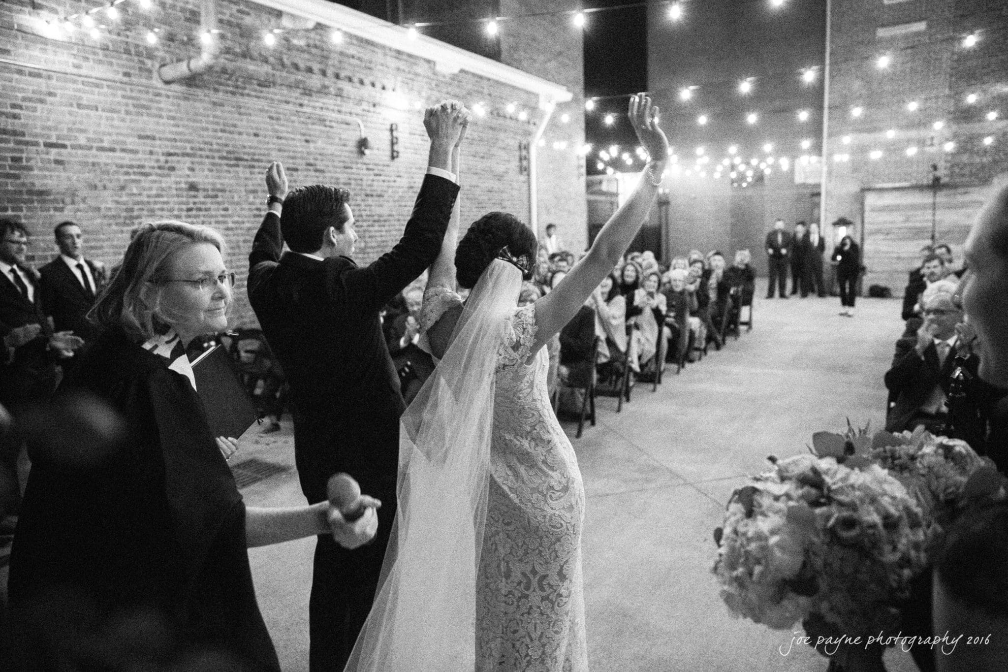 cloth mill at eno river wedding - eleanna & michael A newlywed couple celebrates with raised hands as they exit under string lights during an outdoor evening wedding ceremony. Guests are seated on both sides, and the scene is set against a brick wall backdrop.