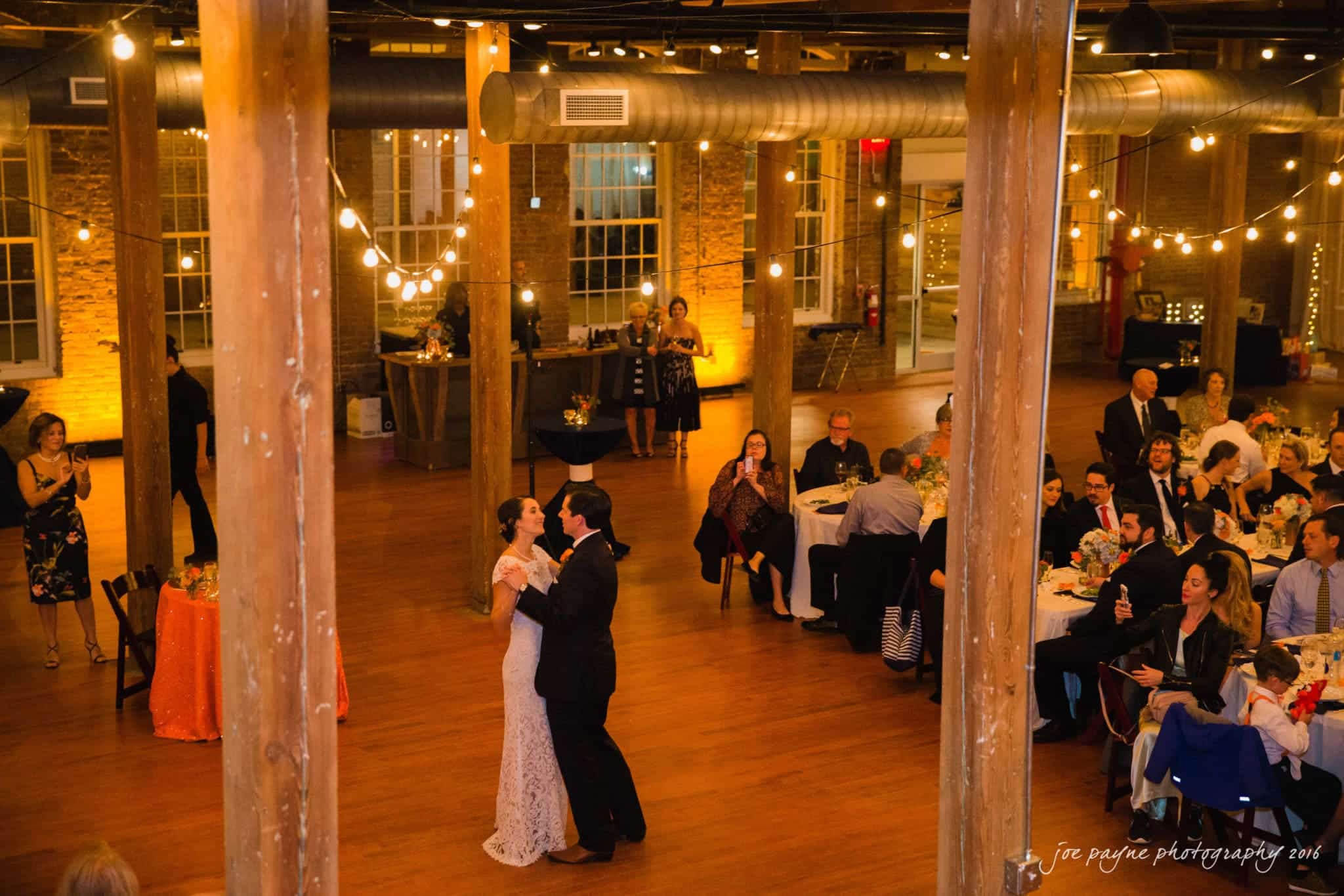 cloth mill at eno river wedding - eleanna & michael A bride and groom share a first dance in a warmly lit, rustic venue with exposed beams. Guests are seated at round tables decorated with white and orange tablecloths, and string lights hang overhead.