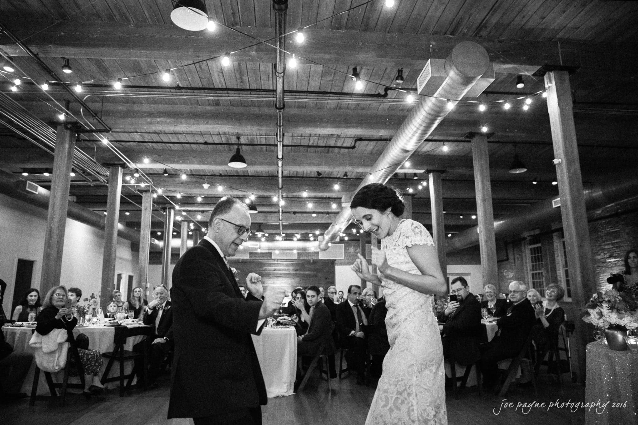 cloth mill at eno river wedding - eleanna & michael A bride and an older man, possibly her father, joyfully dance together at a wedding reception. They are surrounded by seated guests in a warmly lit, rustic venue with wooden beams and string lights. The guests watch and smile at the scene.