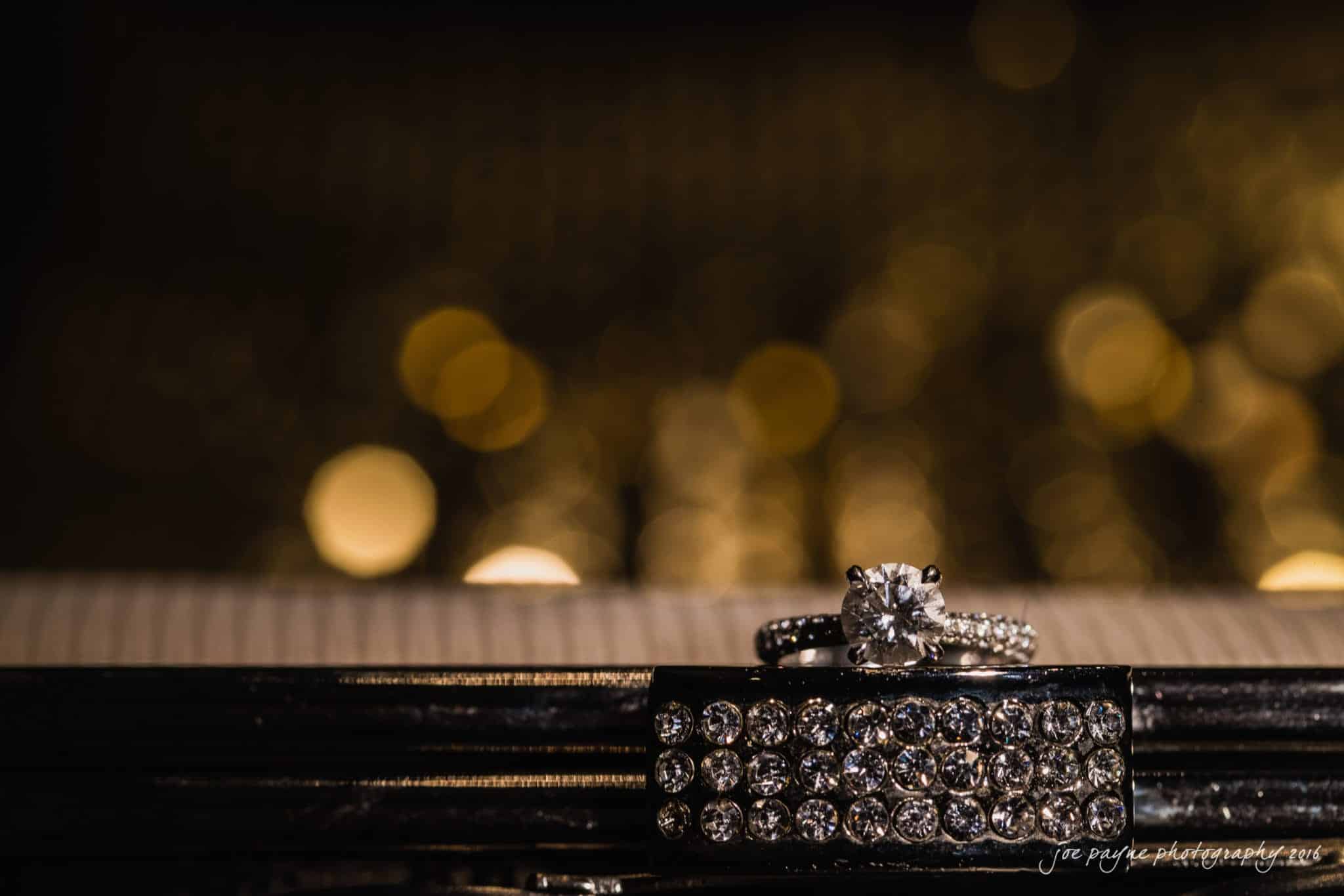 cloth mill at eno river wedding - eleanna & michael A close-up image of a diamond engagement ring and a wedding band with smaller diamonds, resting on a textured, sparkling surface. The background is blurred with warm, golden bokeh lights.