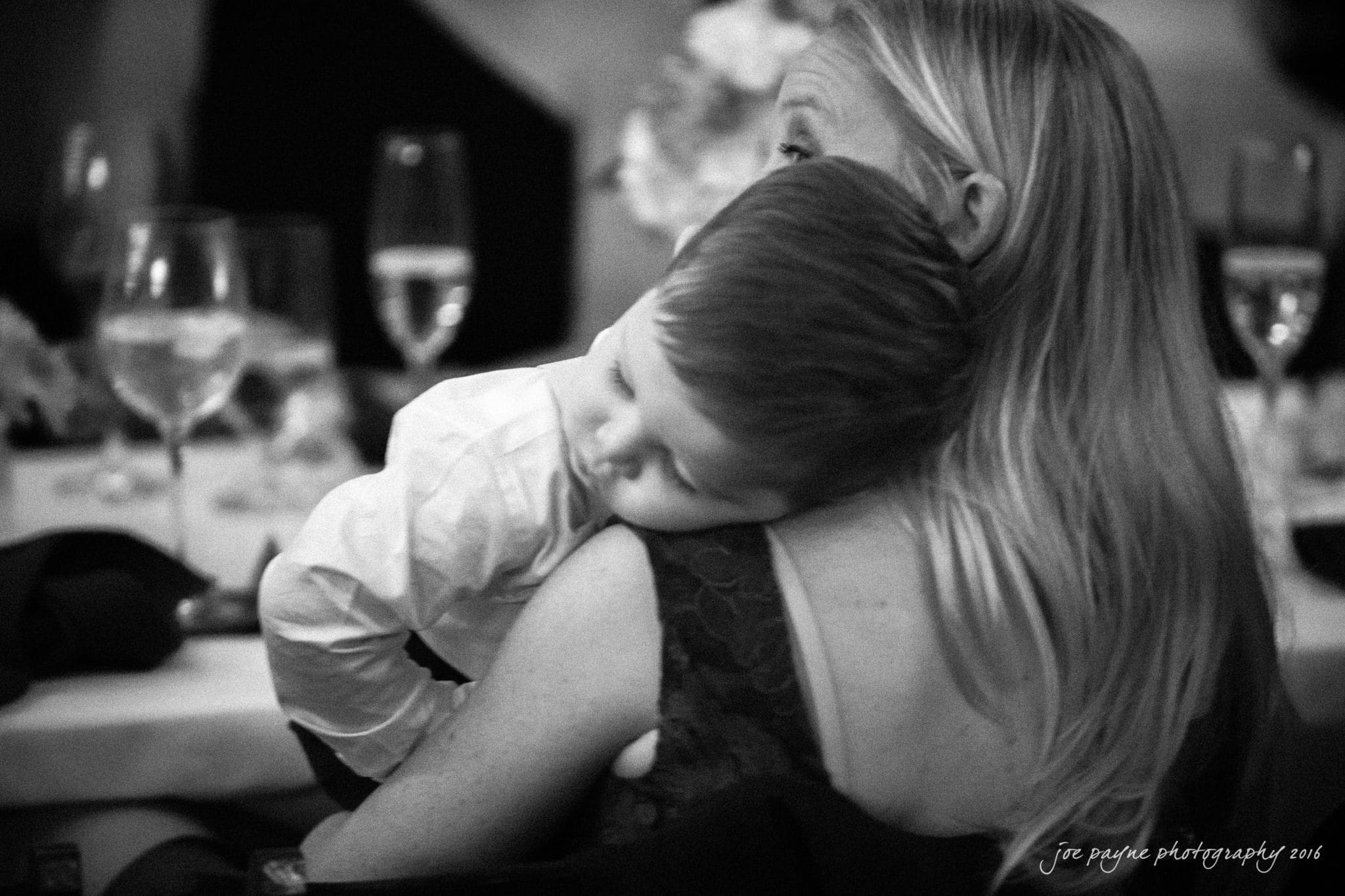 cloth mill at eno river wedding - eleanna & michael A black and white photo of a young child wearing a white shirt sleeping on a womans shoulder. The woman has long, light hair. They are sitting at a table with glasses and blurred background.