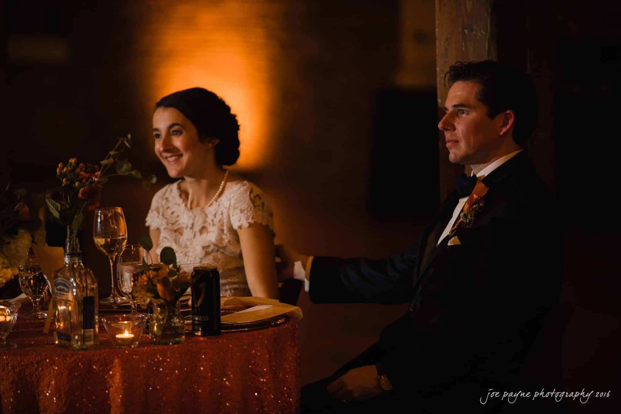 cloth mill at eno river wedding - eleanna & michael A bride and groom sit together at a dimly lit wedding reception. The bride smiles, wearing a white lace dress, while the groom, in a dark suit with an orange boutonniere, gently rests his arm on her chair. The table is adorned with flowers and candles.