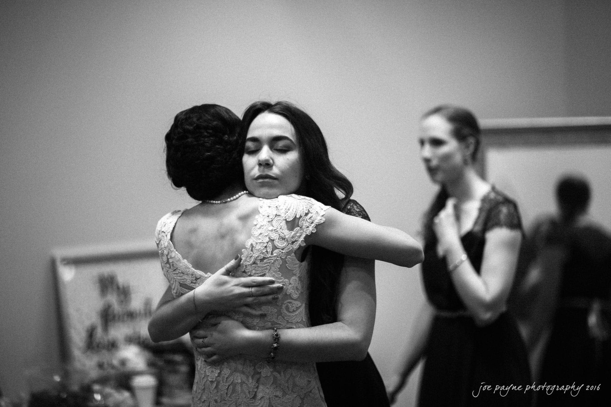 cloth mill at eno river wedding - eleanna & michael A black and white photo of two women embracing warmly, with their eyes closed, in the foreground. Another woman stands in the background, holding her hand to her chest. A framed picture and a table are visible behind them.