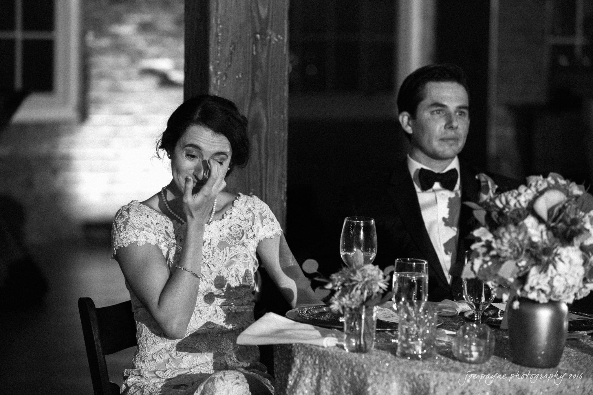 cloth mill at eno river wedding - eleanna & michael A woman in a floral dress sitting at a table wipes her eye, appearing emotional. Next to her, a man in a suit and bow tie looks on. The table is adorned with flowers and glasses. The setting is dimly lit, suggesting an intimate event or celebration.