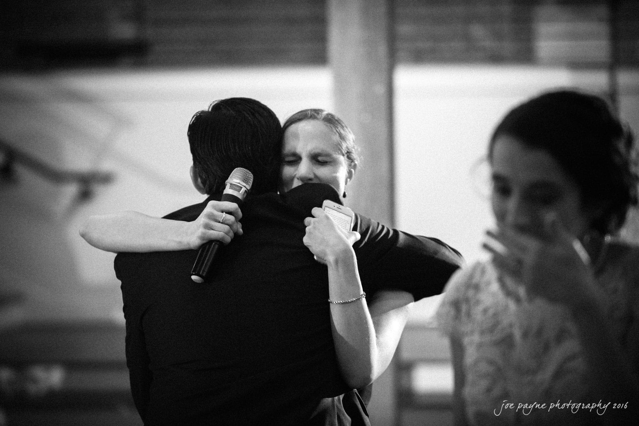 cloth mill at eno river wedding - eleanna & michael A black-and-white photo of a person hugging another who is holding a microphone. The embrace appears emotional. A third person is seen to the side, looking downward with a hand near their face.