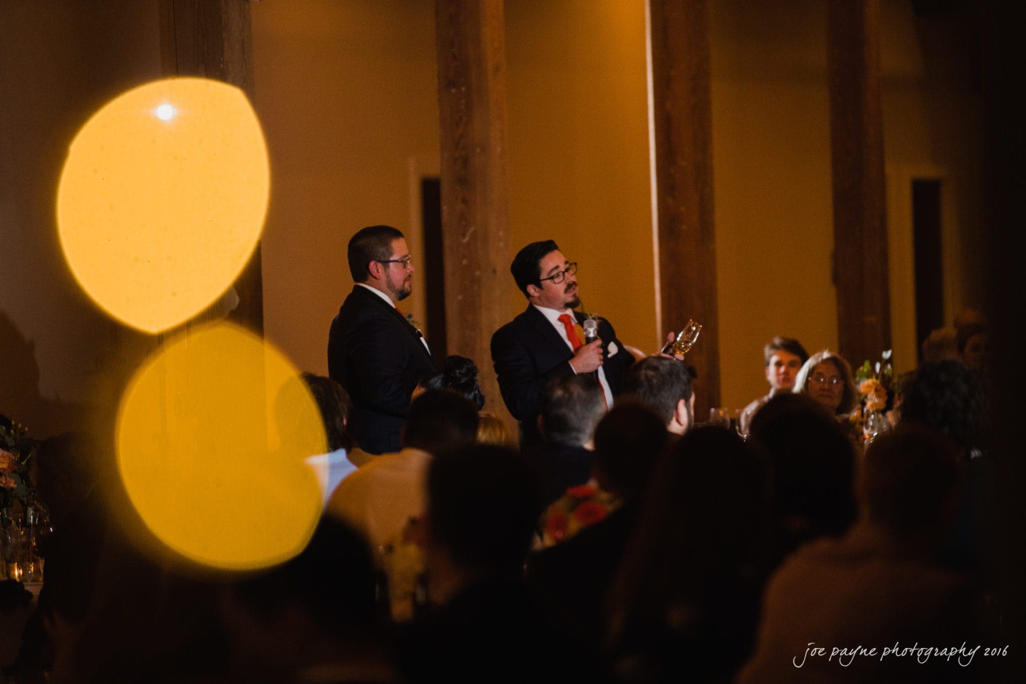cloth mill at eno river wedding - eleanna & michael Two men in suits are standing and speaking into microphones at a formal event. They are surrounded by seated guests. Warm lighting creates a cozy atmosphere, with two blurred, glowing orbs in the foreground.