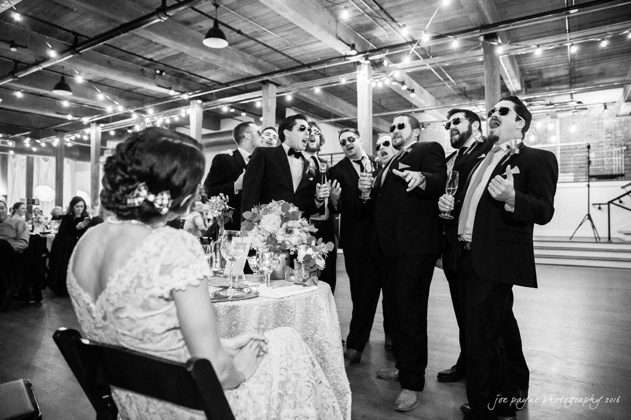cloth mill at eno river wedding - eleanna & michael A group of men in suits and sunglasses sing around a seated woman at a wedding reception. The indoor setting features string lights and wooden beams. The woman, in a lace dress, watches them with amusement.