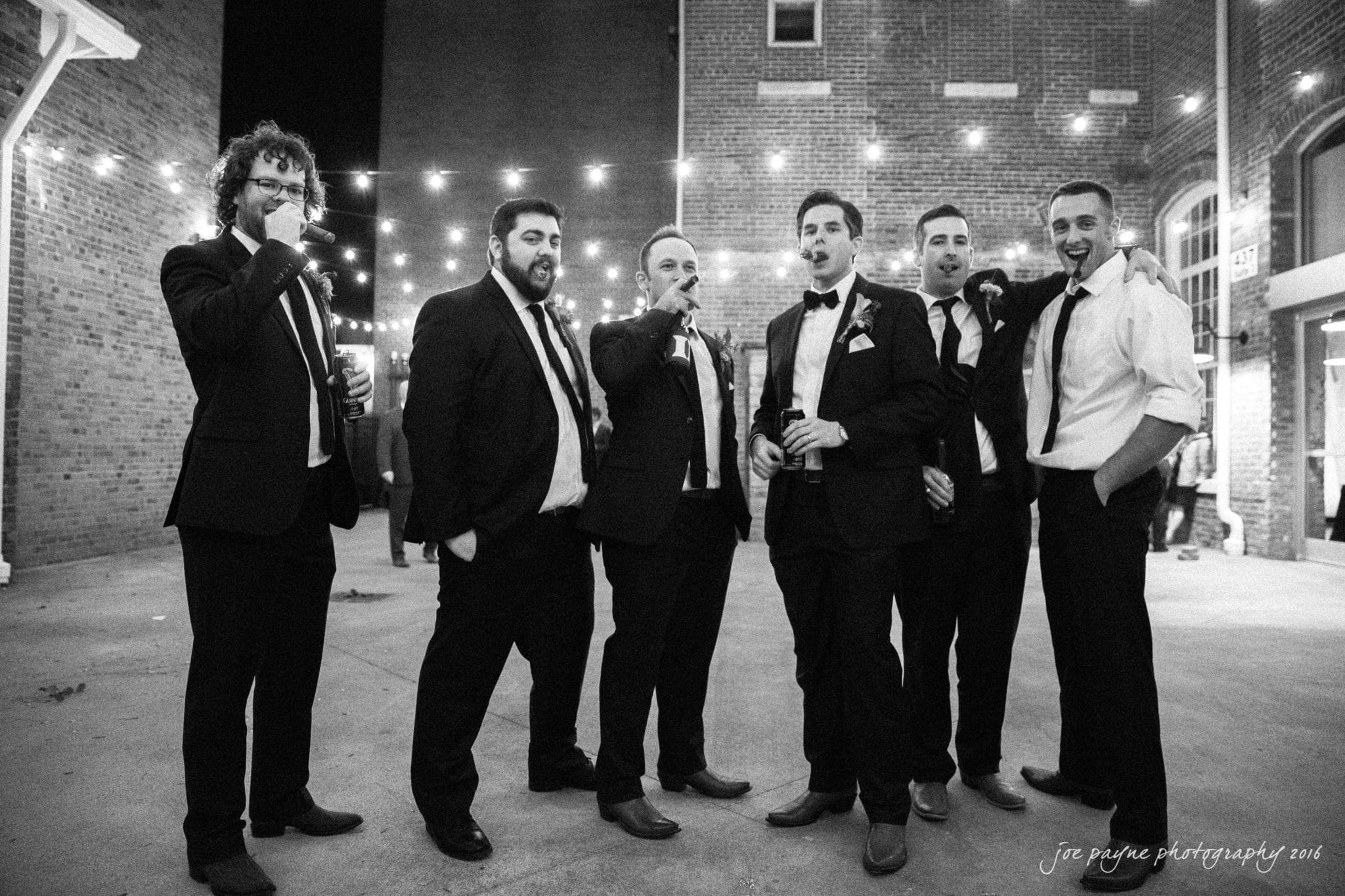 cloth mill at eno river wedding - eleanna & michael A group of six men dressed in formal wear stand outdoors at night, some holding cigars. String lights are overhead, and a brick building is in the background. They appear to be celebrating or posing casually.