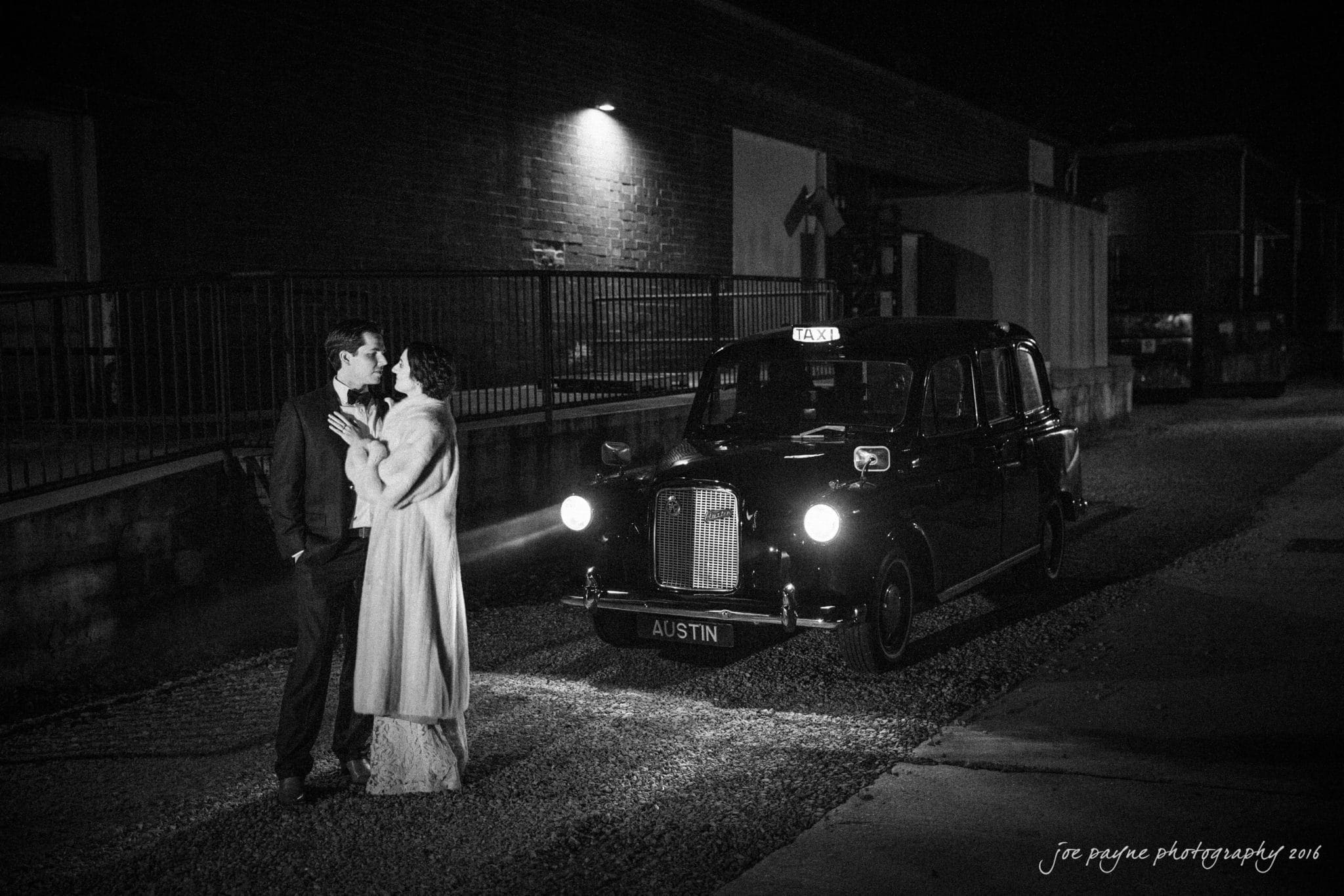 cloth mill at eno river wedding - eleanna & michael A couple stands affectionately beside a classic black taxi in a dimly lit street. The woman wears a long coat and gown, while the man is in a suit. The atmosphere is romantic, with a vintage feel.