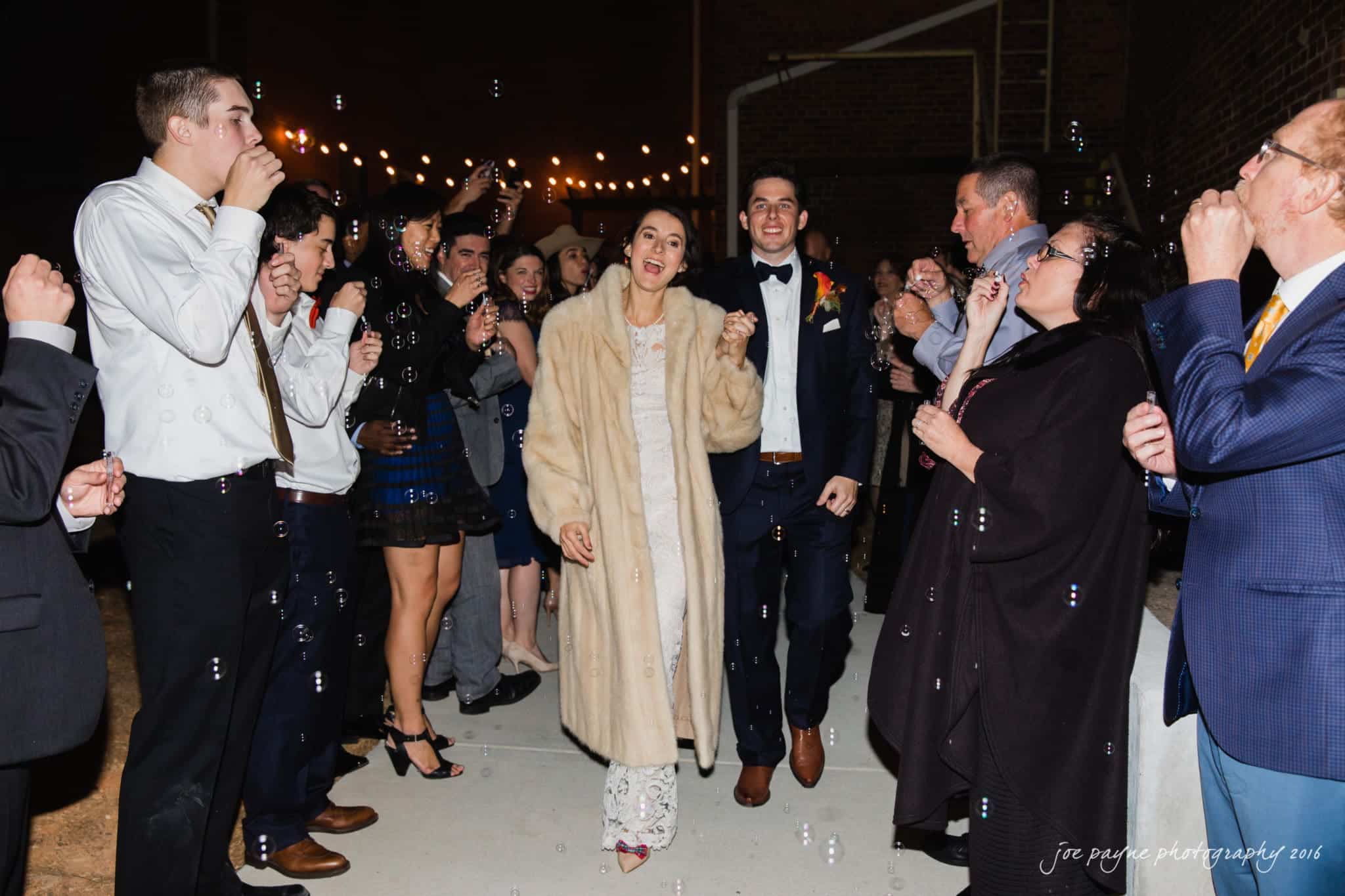 cloth mill at eno river wedding - eleanna & michael A couple in formal attire walks down a path surrounded by cheering guests blowing bubbles. The woman wears a fur coat, and the man is in a dark suit. String lights hang above, creating a festive atmosphere at night.