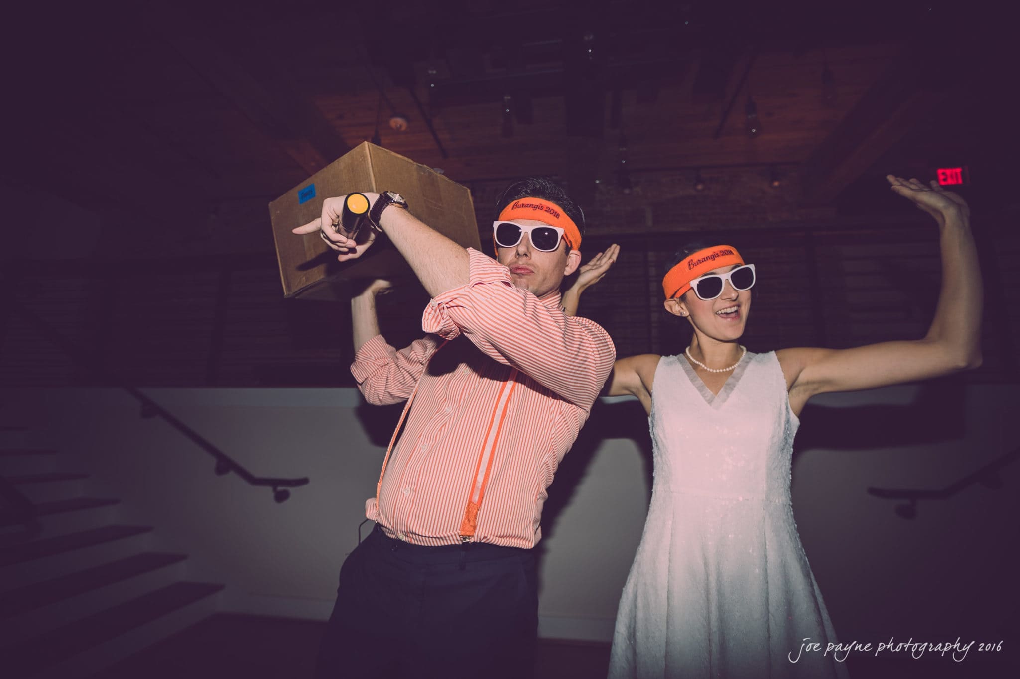 cloth mill at eno river wedding - eleanna & michael A person in a striped shirt with suspenders and a cardboard box poses with a person in a white dress. Both wear matching orange headbands and sunglasses. They appear to be at a lively indoor event, captured in a fun, playful moment.