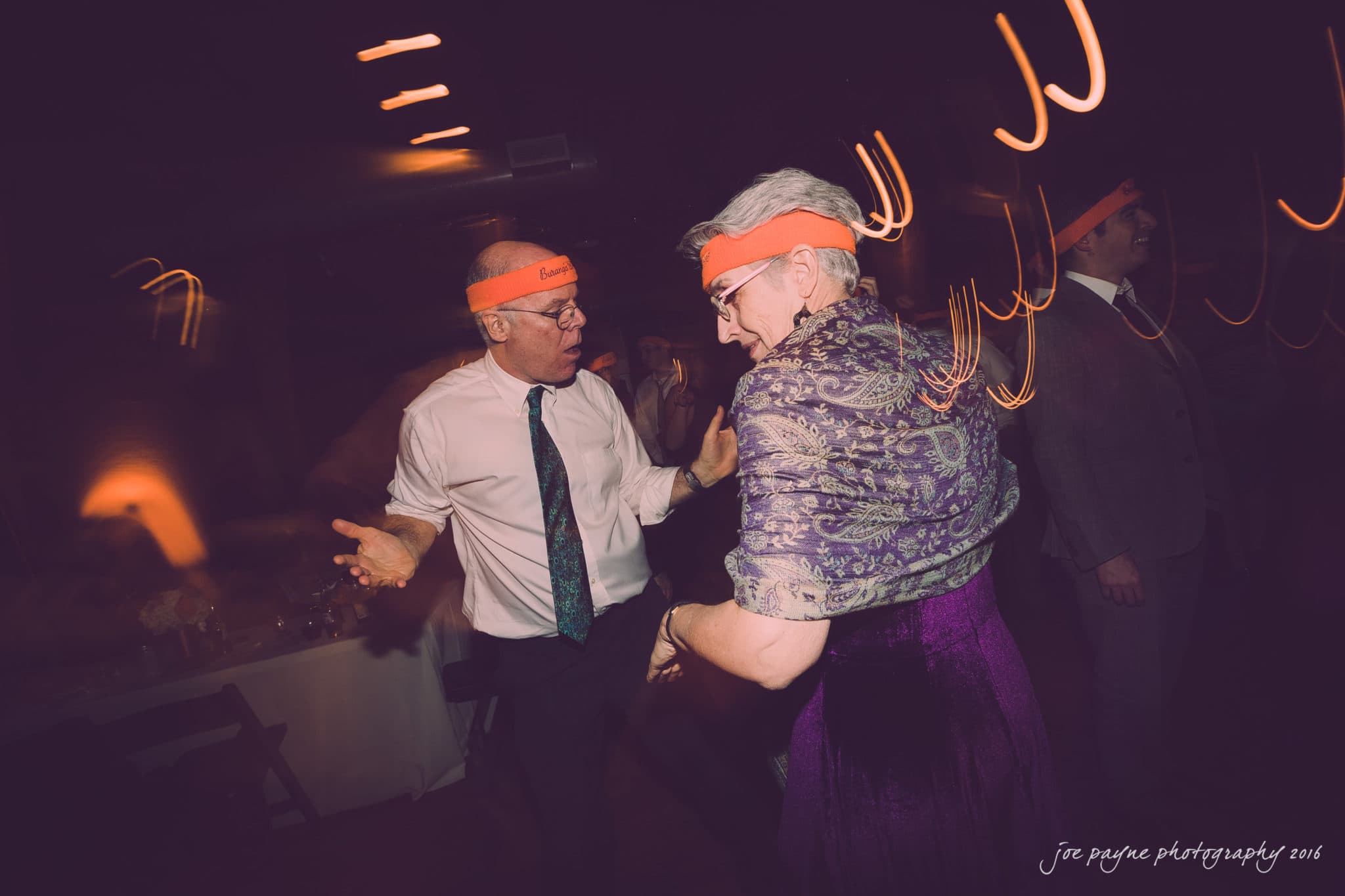 cloth mill at eno river wedding - eleanna & michael A man and a woman wearing orange headbands dance energetically in a dimly lit room. The man is in a white shirt and tie, and the woman wears a purple outfit with a patterned shawl. The image has motion blur, enhancing the lively atmosphere.