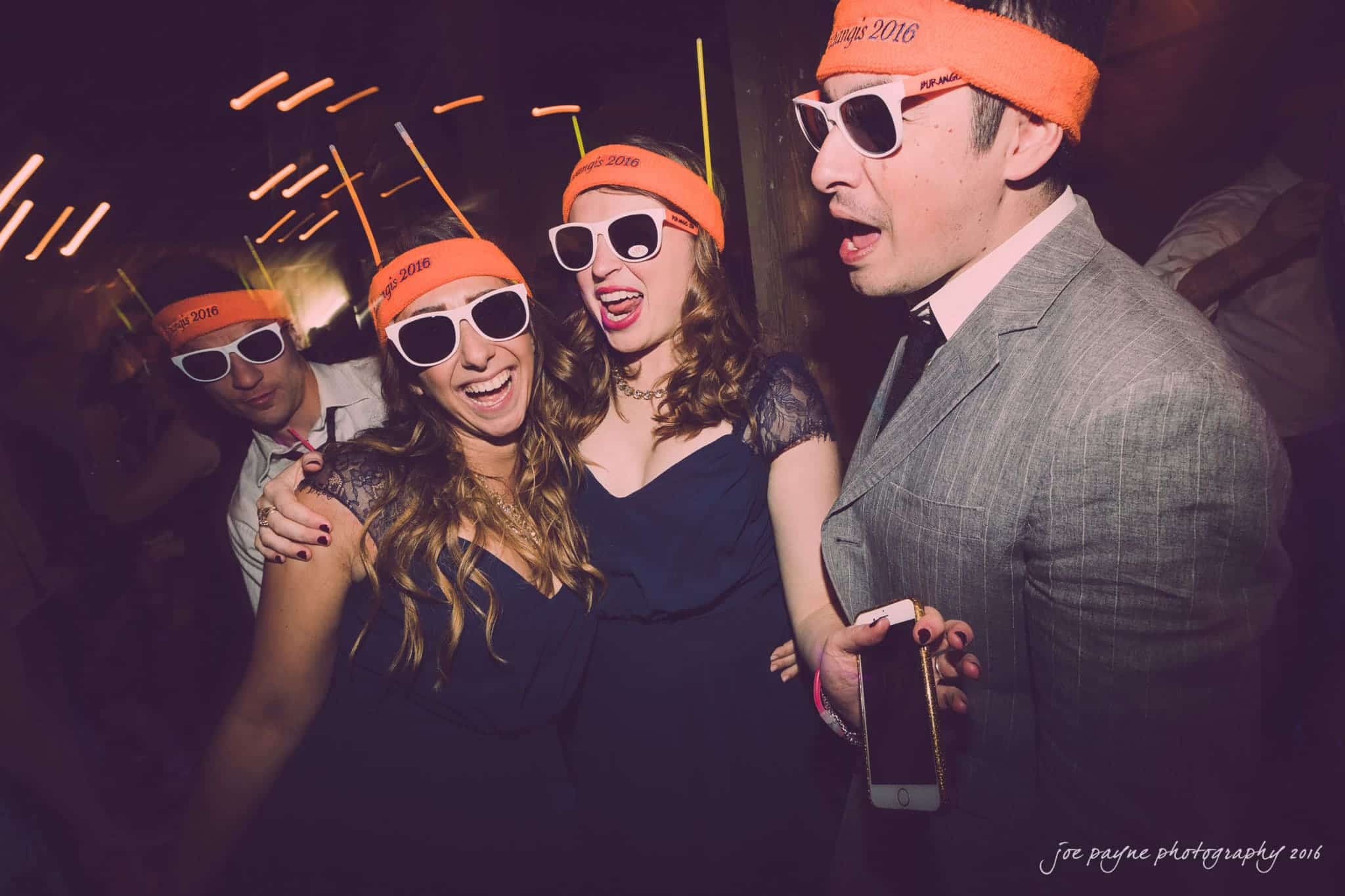 cloth mill at eno river wedding - eleanna & michael Four people at a party, wearing Vegas 2016 orange headbands and sunglasses, laughing and enjoying themselves. One person in a suit holds a smartphone. The background features blurred, dynamic lights.