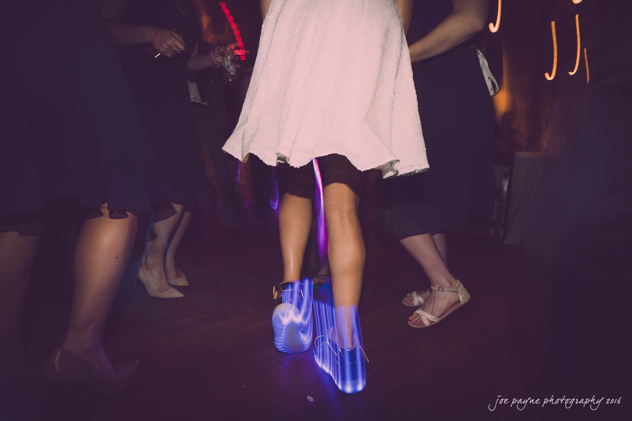 cloth mill at eno river wedding - eleanna & michael People dancing at a party, with a focus on a person in a white dress wearing shoes that glow with blue light. Others in dark clothing are visible in the background.