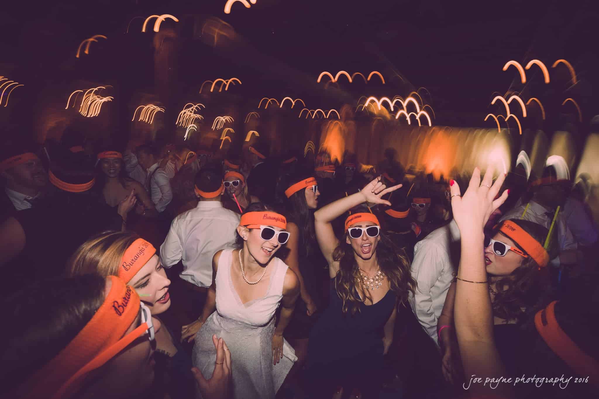 cloth mill at eno river wedding - eleanna & michael A lively dance floor at a wedding party, with people wearing matching orange headbands and white sunglasses. The lighting creates a dynamic atmosphere with motion blurs and light trails, adding to the energetic vibe.