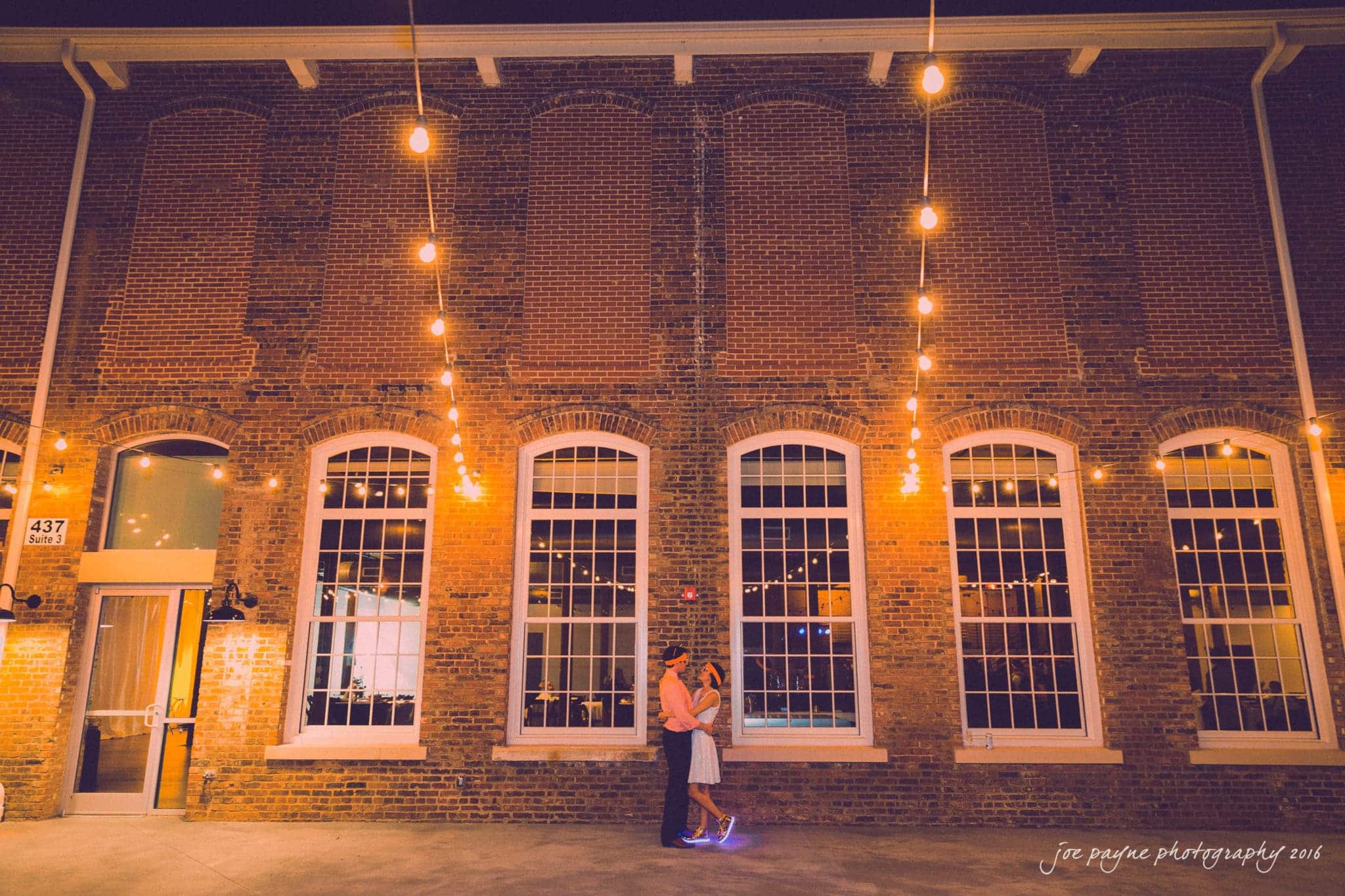 cloth mill at eno river wedding - eleanna & michael A couple embraces under a string of warm lights against a brick building with large windows at night. The setting has a cozy, romantic ambiance.