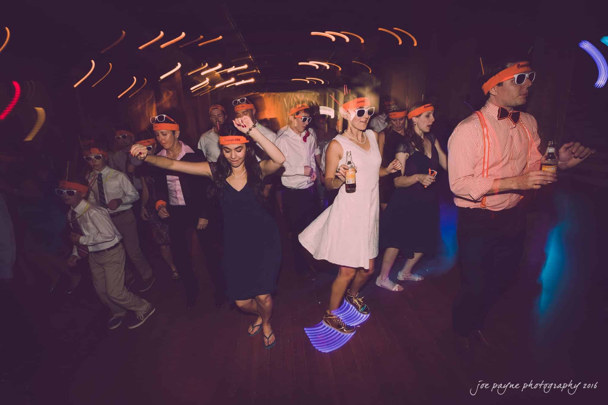 cloth mill at eno river wedding - eleanna & michael A lively group of people dance at a party, wearing bright orange headbands and sunglasses. The room is dimly lit with colorful streaks of light creating a vibrant atmosphere. Some hold drinks as they enjoy the music and festivities.