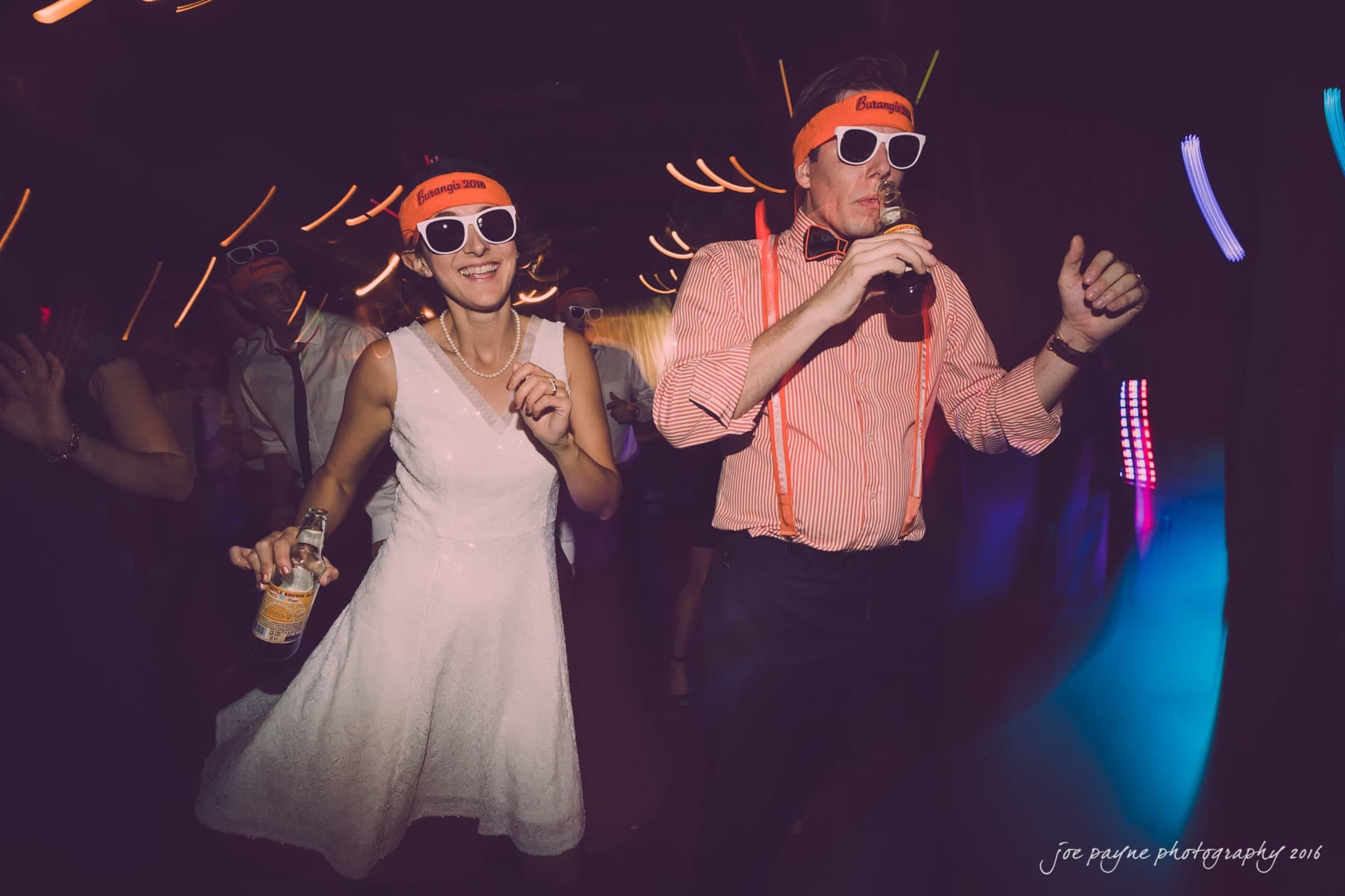 cloth mill at eno river wedding - eleanna & michael A couple wearing matching neon hats and sunglasses dances energetically at a party. The woman is in a white dress, holding a drink, while the man wears a striped shirt with suspenders, also holding a drink. Colorful lights illuminate the scene.
