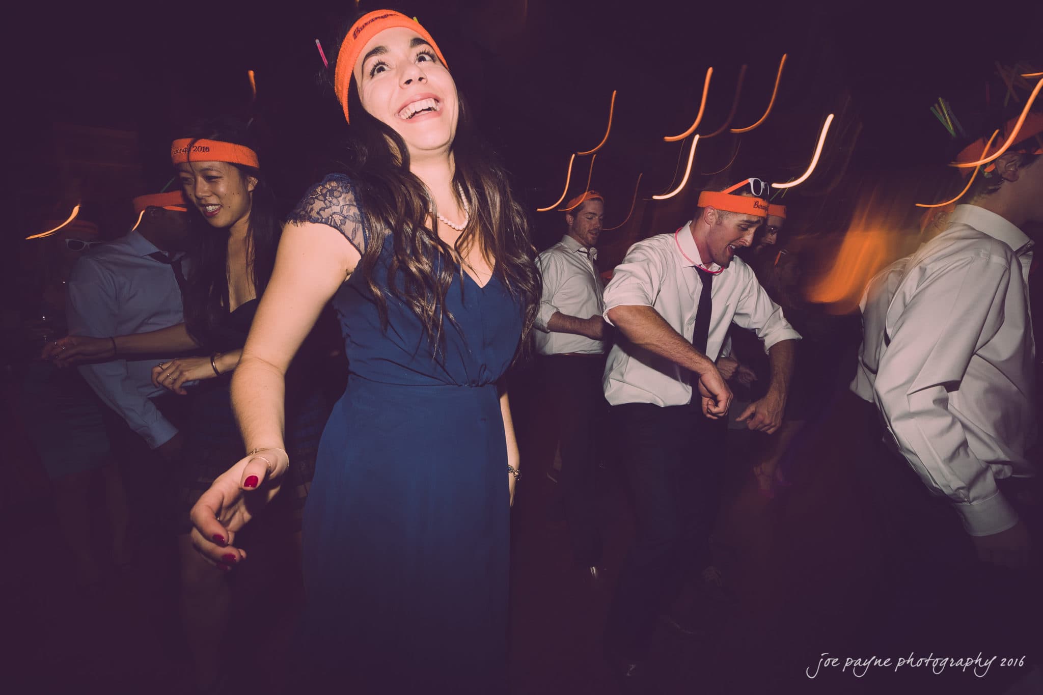 cloth mill at eno river wedding - eleanna & michael A group of people in party attire dance energetically with orange headbands. The woman in the foreground is wearing a navy blue dress, smiling broadly. The background shows blurred motion, capturing the lively atmosphere.