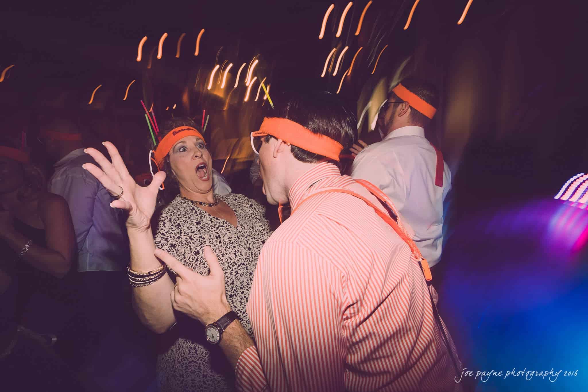 cloth mill at eno river wedding - eleanna & michael A man and woman wearing orange headbands energetically dance at a party. The woman looks surprised, with her hands up. Blurred lights and people in the background create a lively atmosphere.