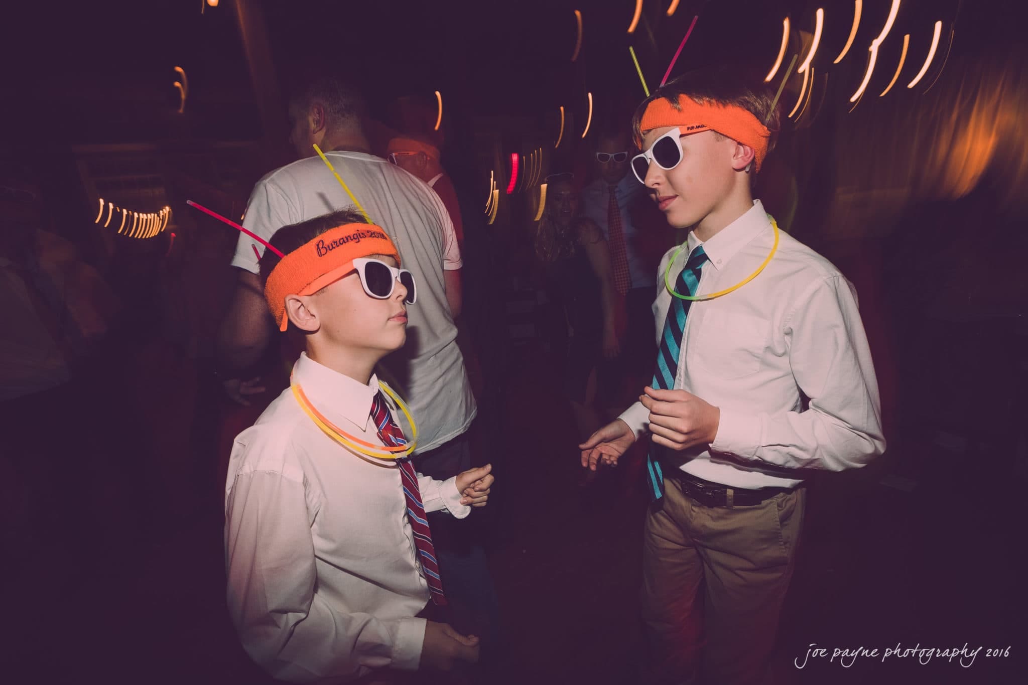 cloth mill at eno river wedding - eleanna & michael Two boys wearing sunglasses, orange headbands with straws, and glow necklaces dance in a dimly lit room. They are dressed in white shirts and ties. The scene has a festive, party atmosphere with blurred lights in the background.