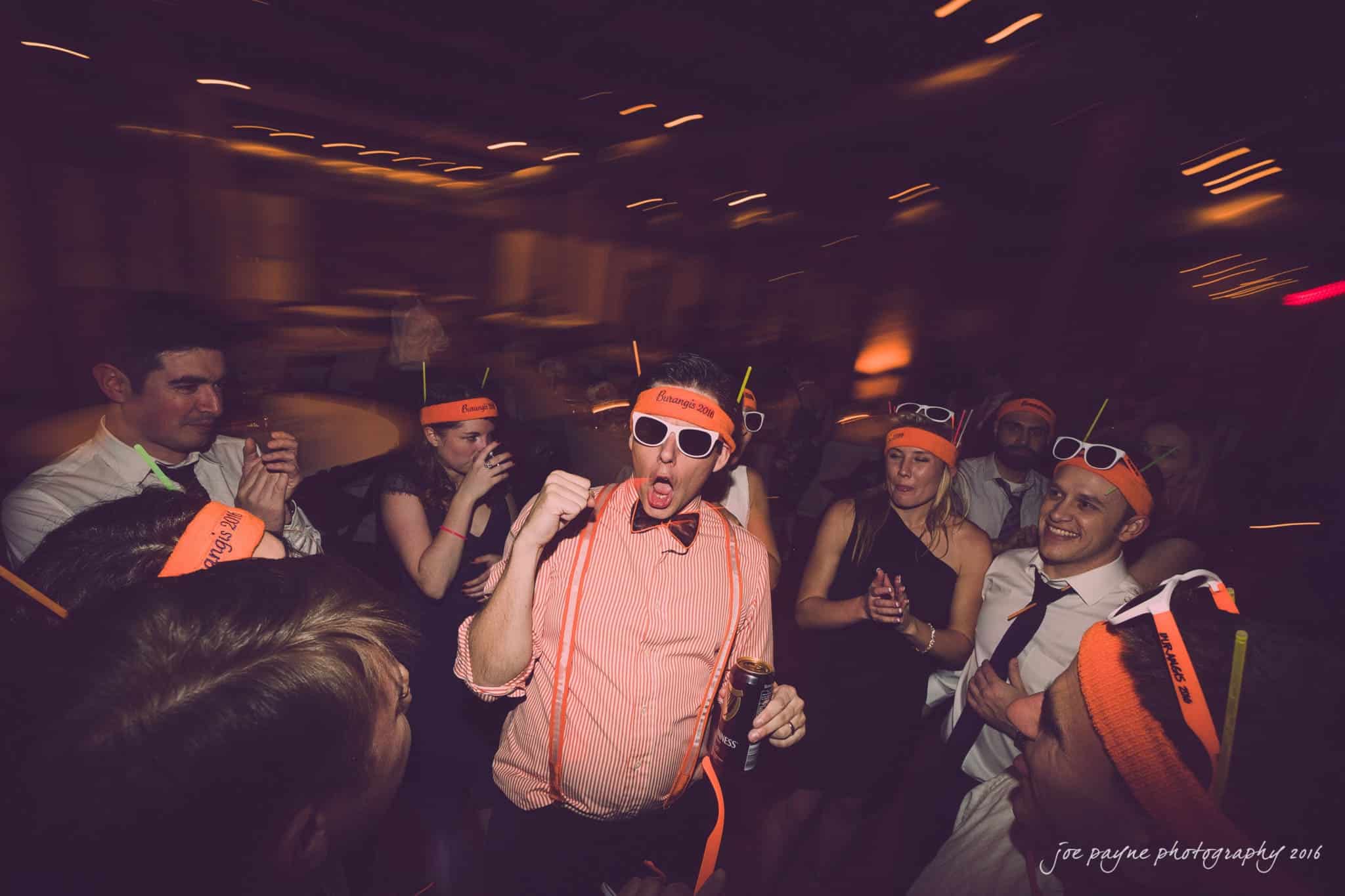 cloth mill at eno river wedding - eleanna & michael A lively party scene with people wearing orange headbands and sunglasses, gathered around a man who is enthusiastically singing or speaking. The ambient lighting creates a festive atmosphere, and everyone appears to be enjoying themselves.