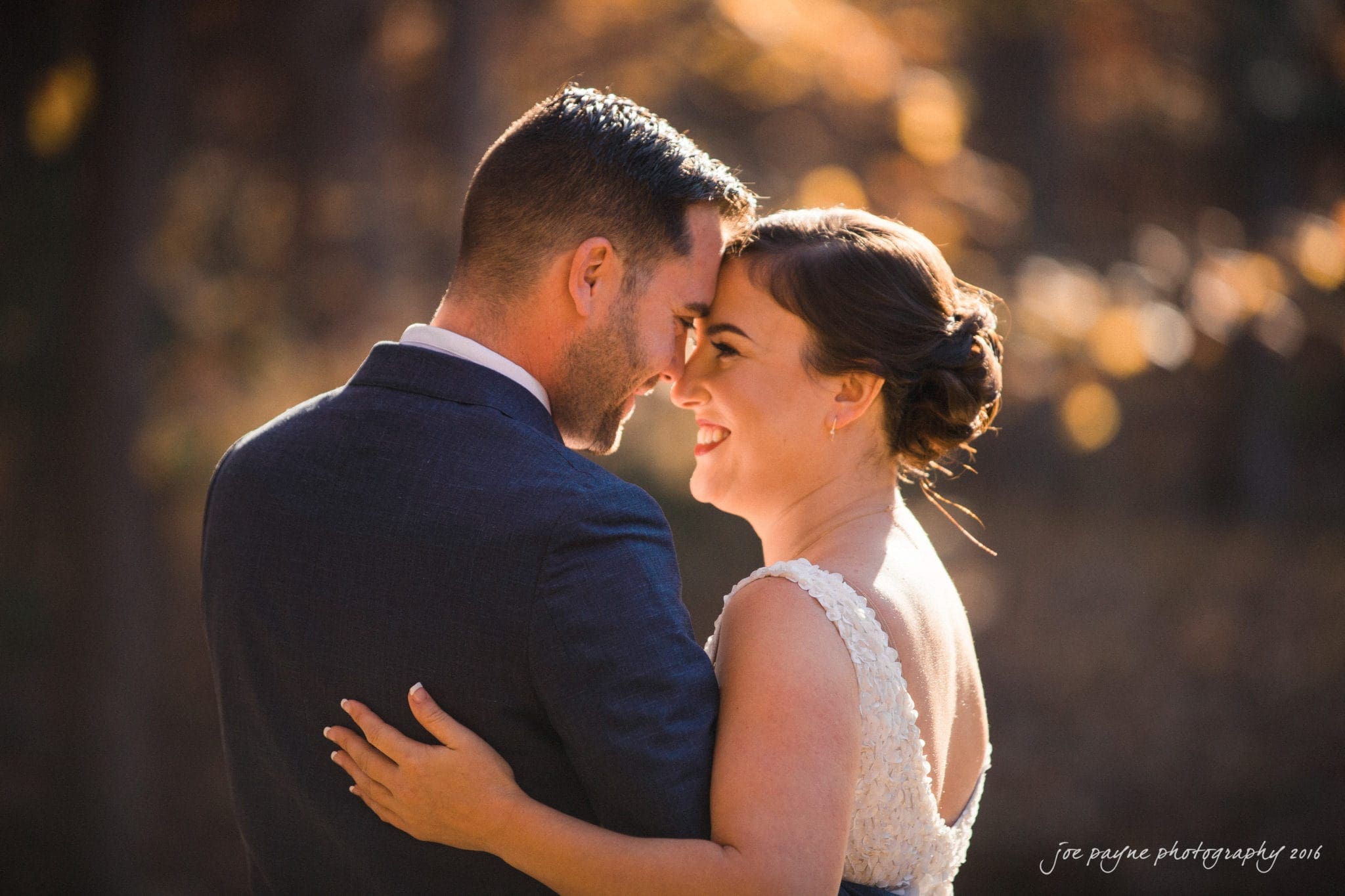 A couple stands close, smiling at each other in an outdoor setting. They are dressed formally, with the woman in a white dress and the man in a dark suit. Sunlight filters through the trees, creating a warm, intimate atmosphere.