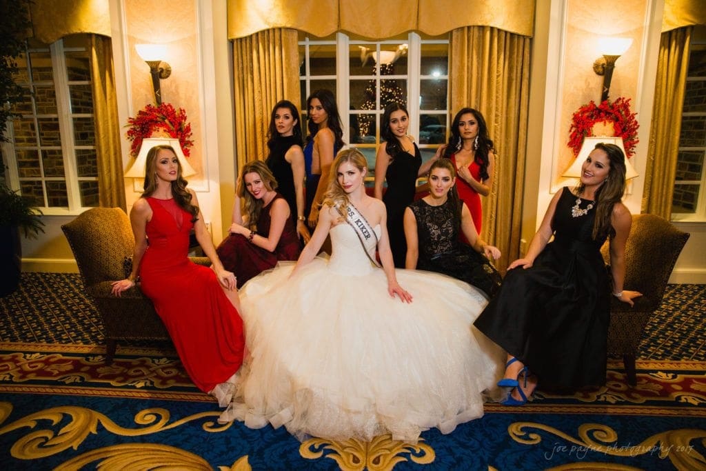 A group of elegantly dressed women in formal gowns pose in a luxurious room with large windows and decorative lighting. The woman in the center wears a white ball gown and a sash. The setting includes ornate carpeting and festive floral arrangements.