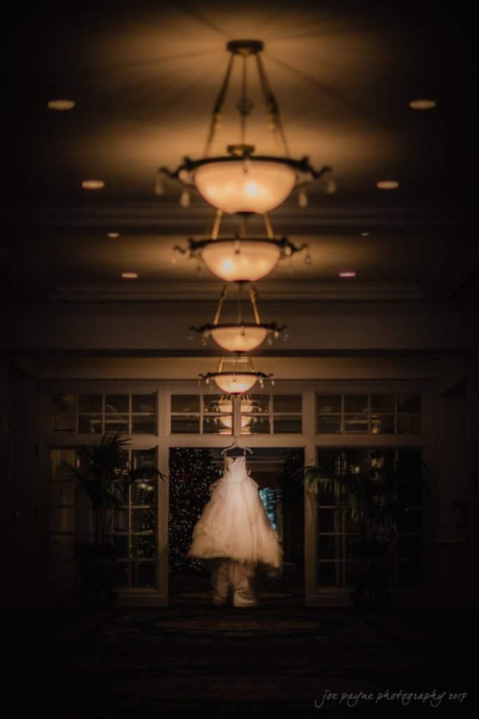 A child in a fluffy white dress runs down a dimly lit hallway, with elegant chandeliers above and a festive, illuminated tree visible through glass doors at the end.