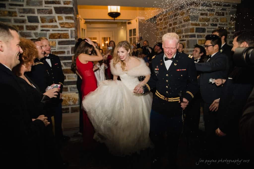 Bride and groom exiting a building while guests celebrate. The bride, in a white gown, and the groom, in military attire, are surrounded by people in formal wear. Guests throw confetti as the couple smiles and holds hands.