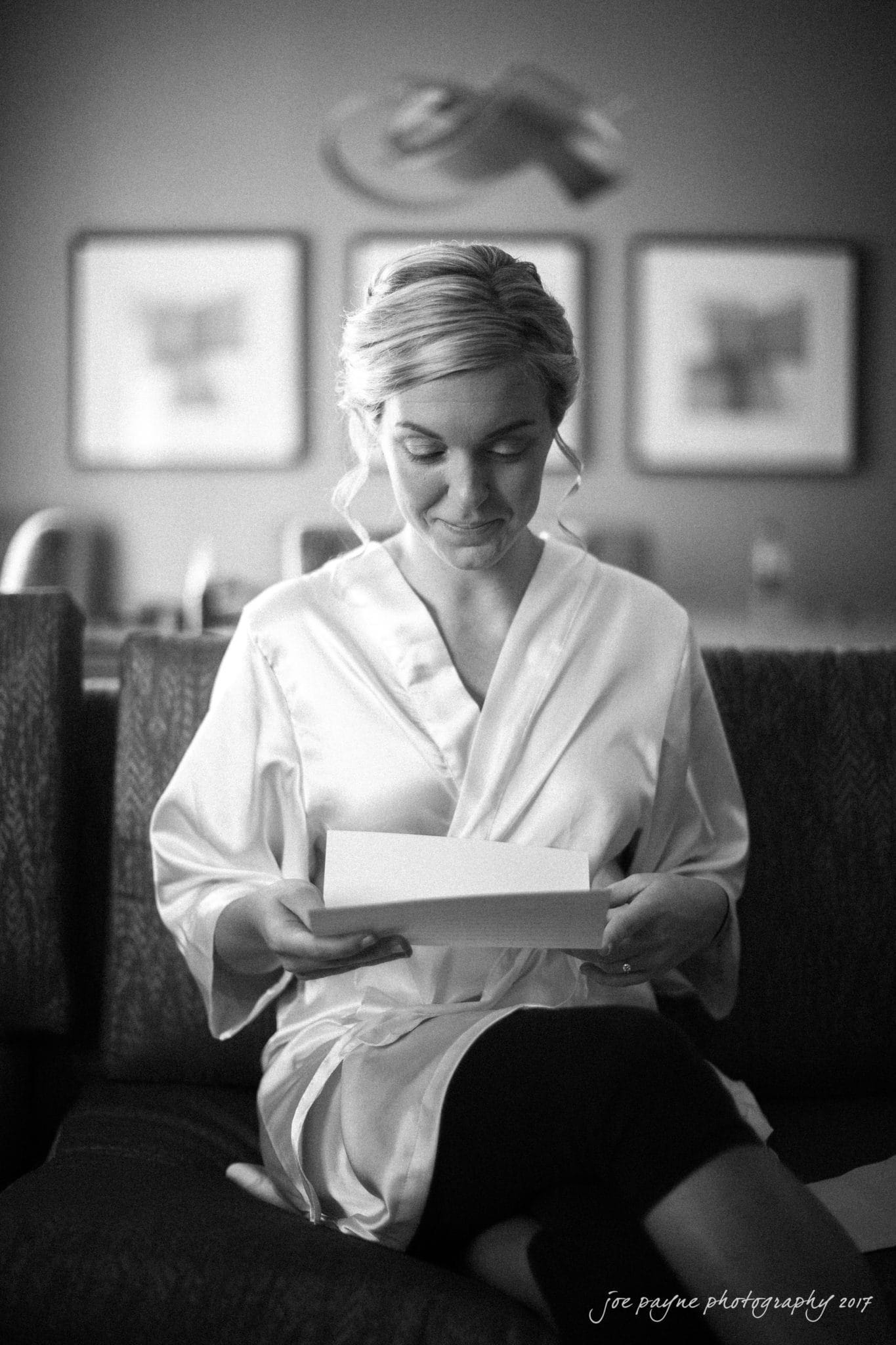 Black and white image of a woman in a silk robe sitting on a sofa, smiling as she reads a card. Three framed artworks are blurred in the background.
