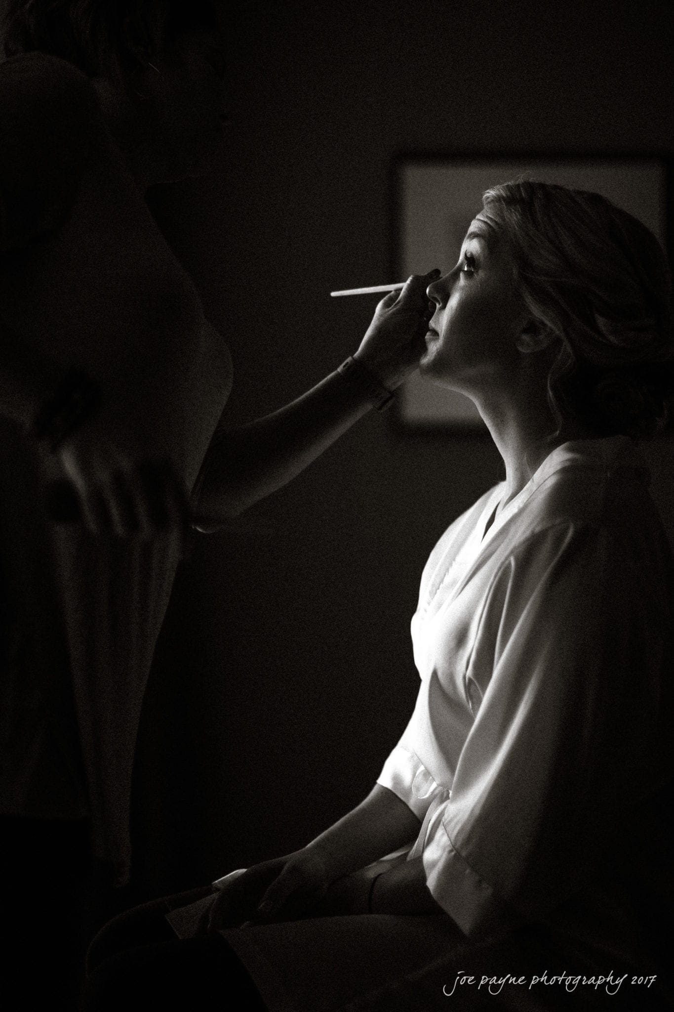 A woman sits in profile, wearing a robe, while having her makeup applied in a dimly lit room. The scene is captured in black and white, creating a dramatic, intimate atmosphere.