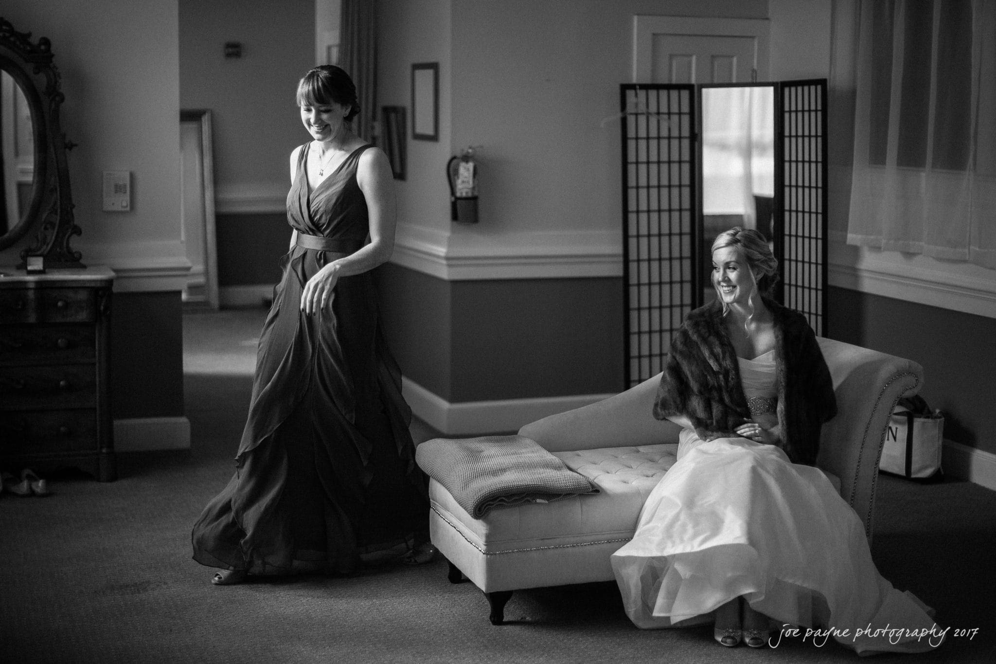 A woman in an elegant gown stands smiling beside another woman seated on a chaise lounge, who is wearing a strapless dress and a shawl. They are in a softly lit room with a large mirror and a decorative folding screen.