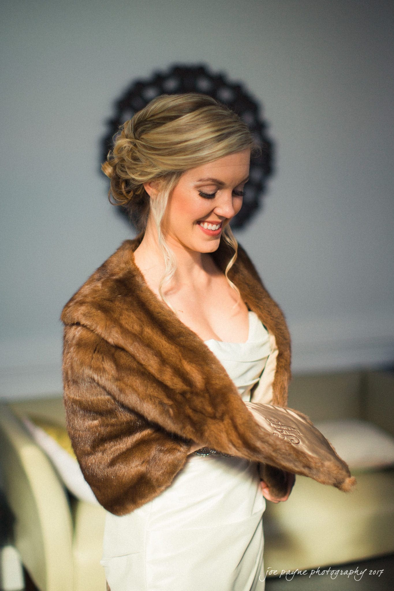 A woman with blonde hair in an elegant updo smiles while wearing an ivory dress and a brown faux fur stole. She is standing indoors with a soft-focus background.