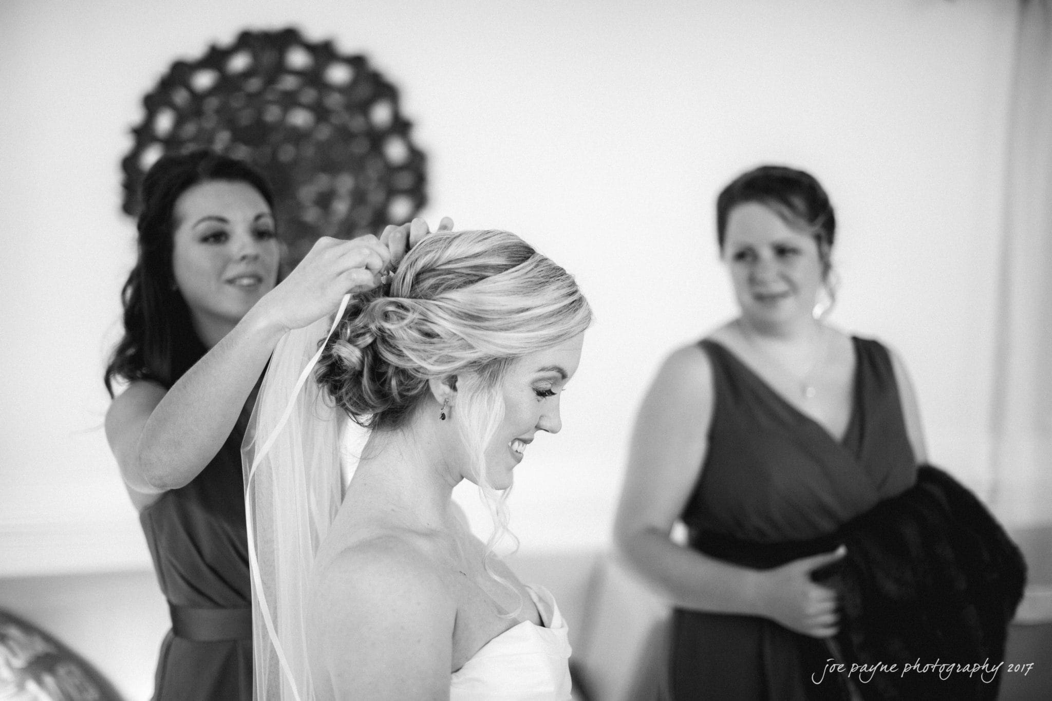 A bride smiles as a woman adjusts her veil, with another woman watching nearby. They are all dressed in formal attire. The room has elegant decor with a decorative wall piece in the background.