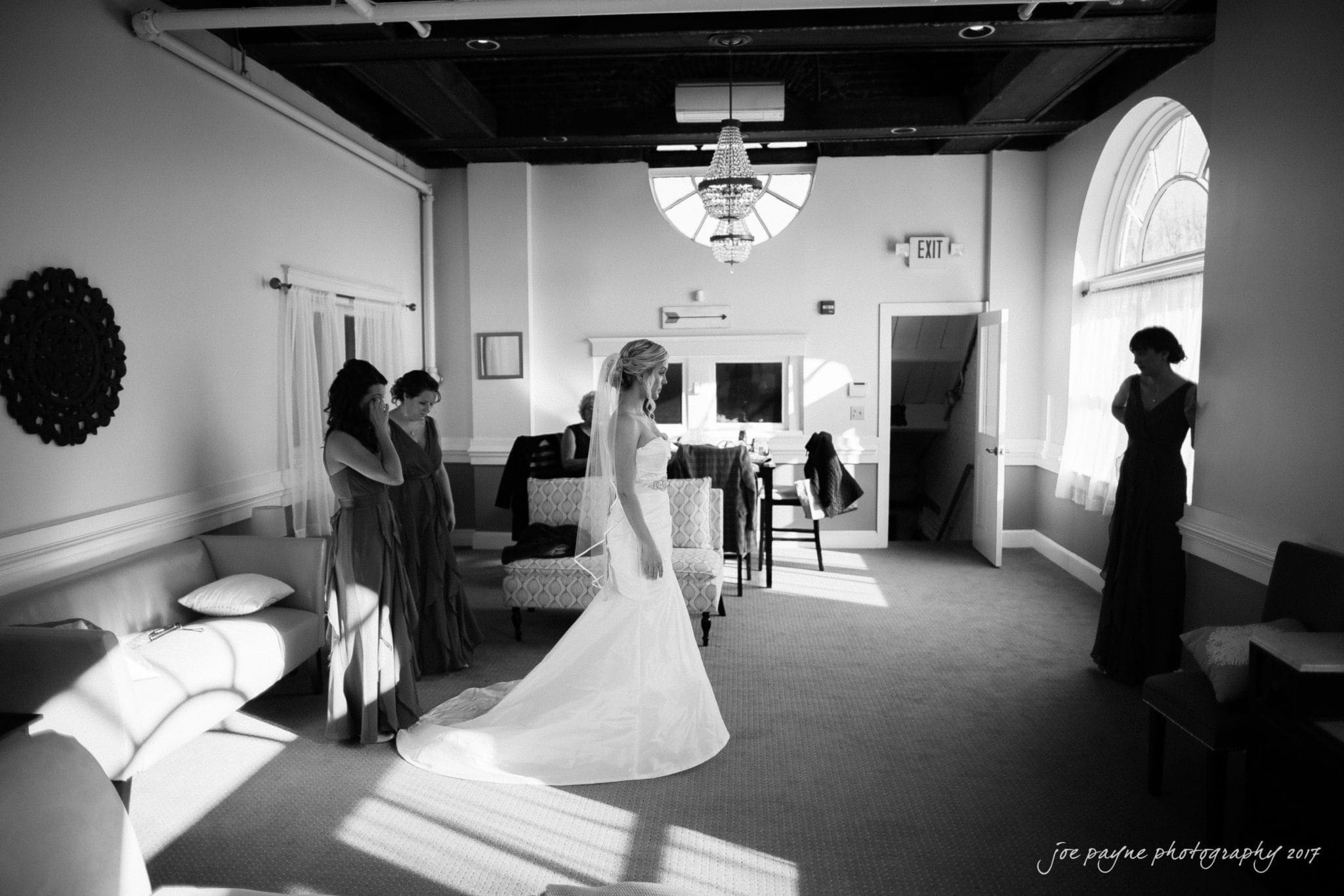 A black and white image of a bride in a strapless gown, illuminated by window light. She stands in a tastefully decorated room with three bridesmaids nearby, lost in quiet preparation moments before the ceremony.