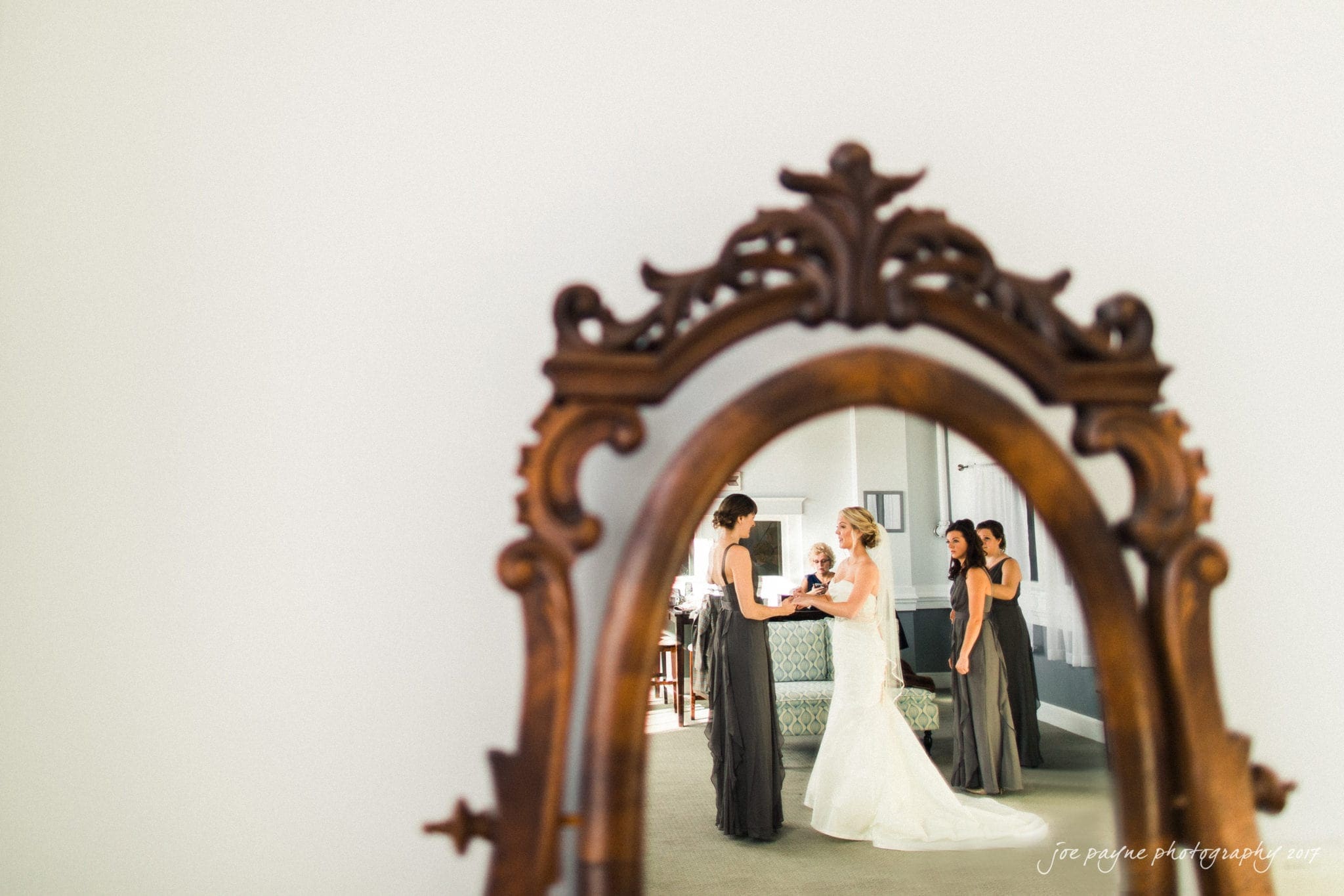 A bride wearing a white gown is seen in a mirror, surrounded by bridesmaids in dark dresses, preparing in a softly lit room. The ornate wooden mirror frame adds a classic touch to the scene.