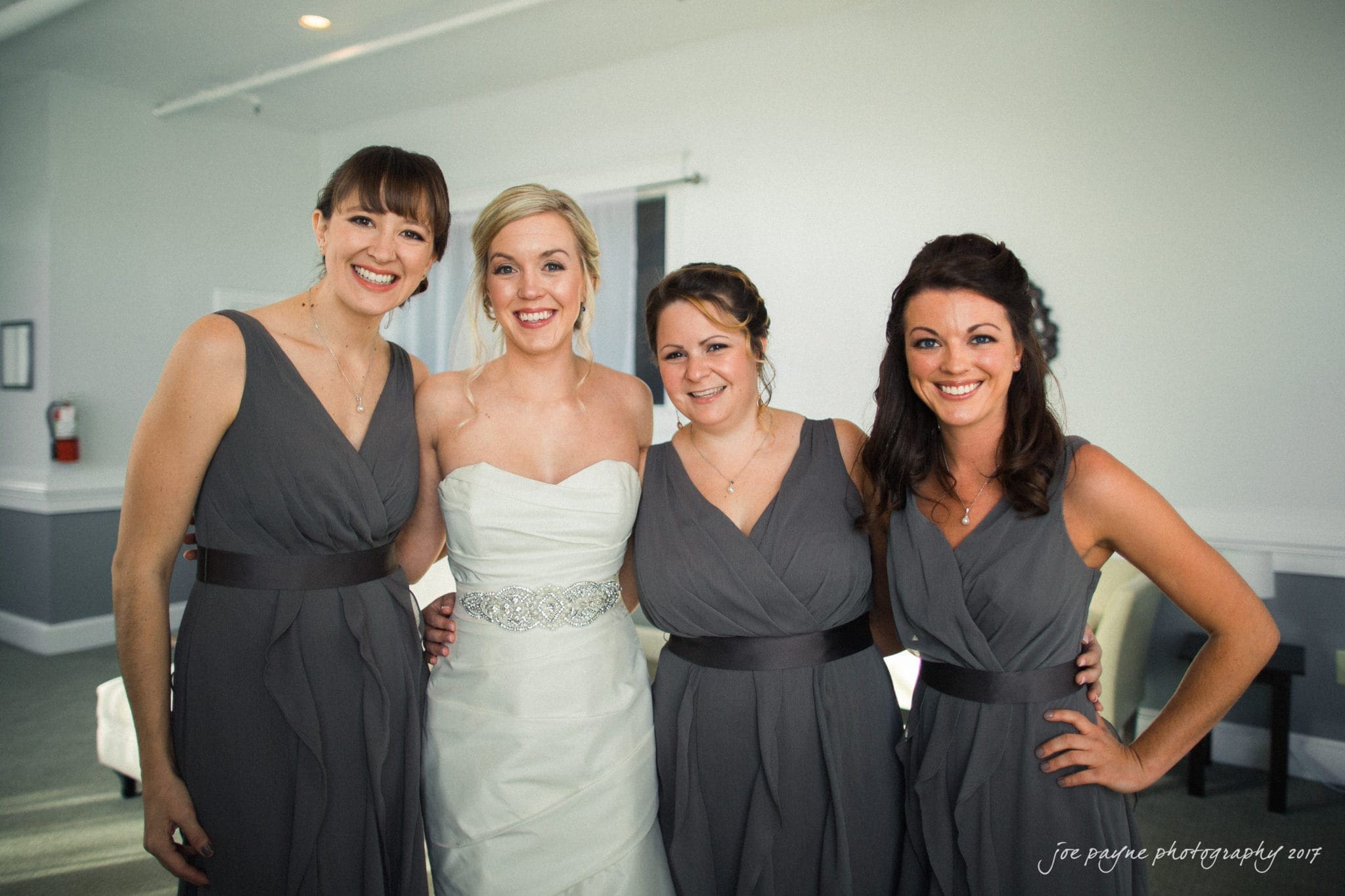 A bride in a white gown smiles while posing with three bridesmaids in matching gray dresses. They stand together in a brightly lit room with light blue walls.