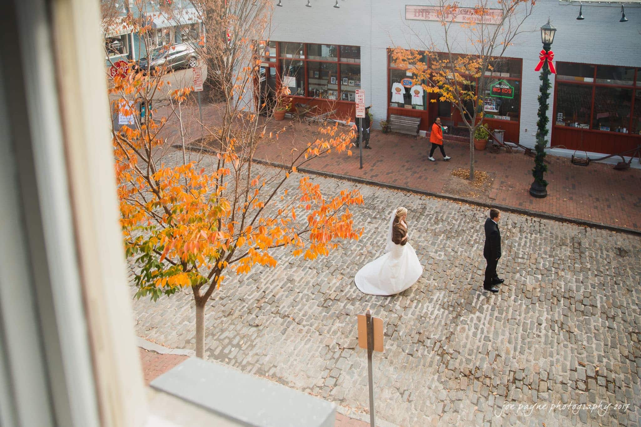 A bride and groom, dressed formally, walk toward each other on a cobblestone street lined with autumn trees. A red bow decorates a lamppost, and a passerby in an orange coat walks by a shopfront in the background.