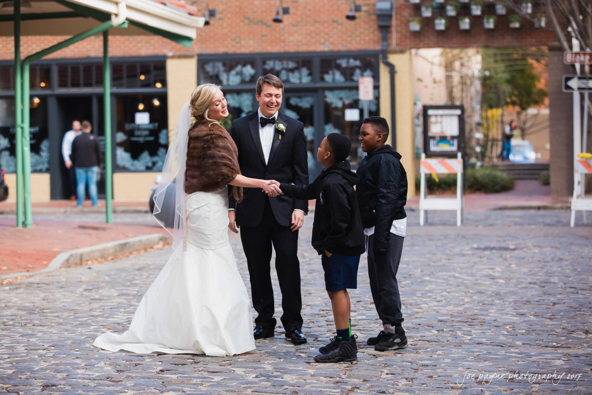 A bride and groom stand on a cobblestone street, smiling and shaking hands with two young boys. The bride wears a white gown and fur shawl, and the groom is in a black tuxedo. The scene appears friendly and candid, set in an urban area.