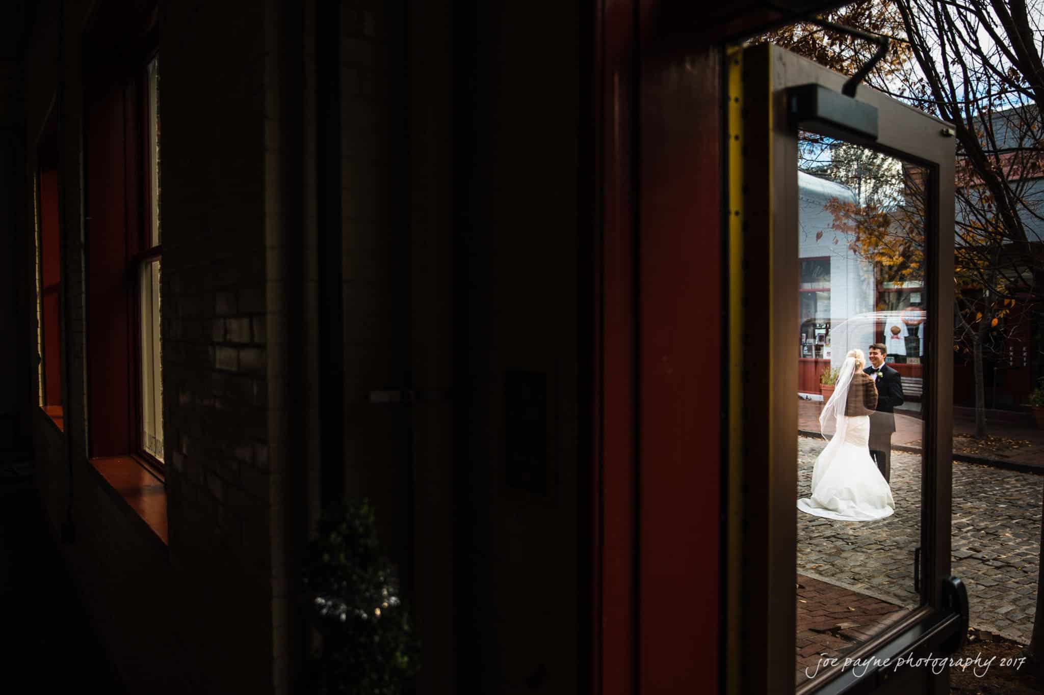 A bride and groom stand outside on a cobblestone path, framed by an open door. The bride wears a white dress and veil, while the groom is in a black suit. Sunlight highlights them against the darker interior.