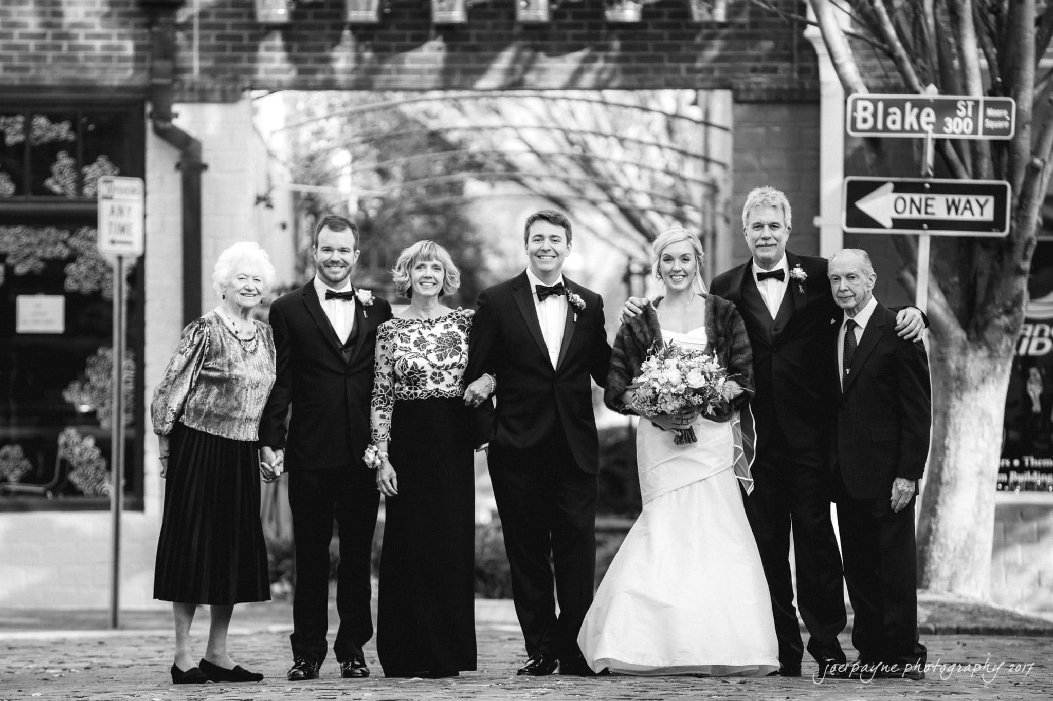 A black-and-white photo of a wedding party standing on a cobblestone street. The group includes a bride in a gown holding a bouquet, a groom in a tuxedo, and four other formally dressed individuals smiling. A street sign and brick archway are in the background.