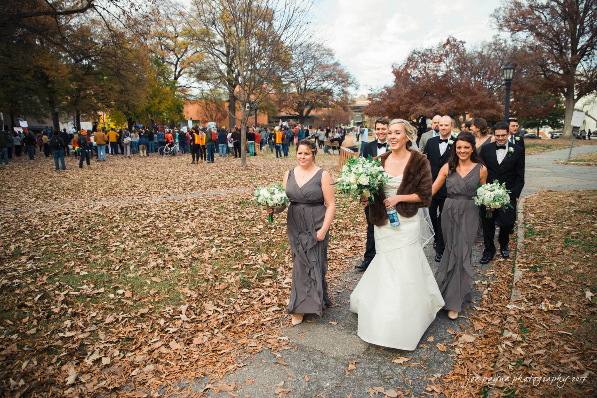 A bride in a white dress and fur stole walks with three bridesmaids in gray dresses and groomsmen in black suits through a park covered in autumn leaves. A crowd is visible in the background among the trees.