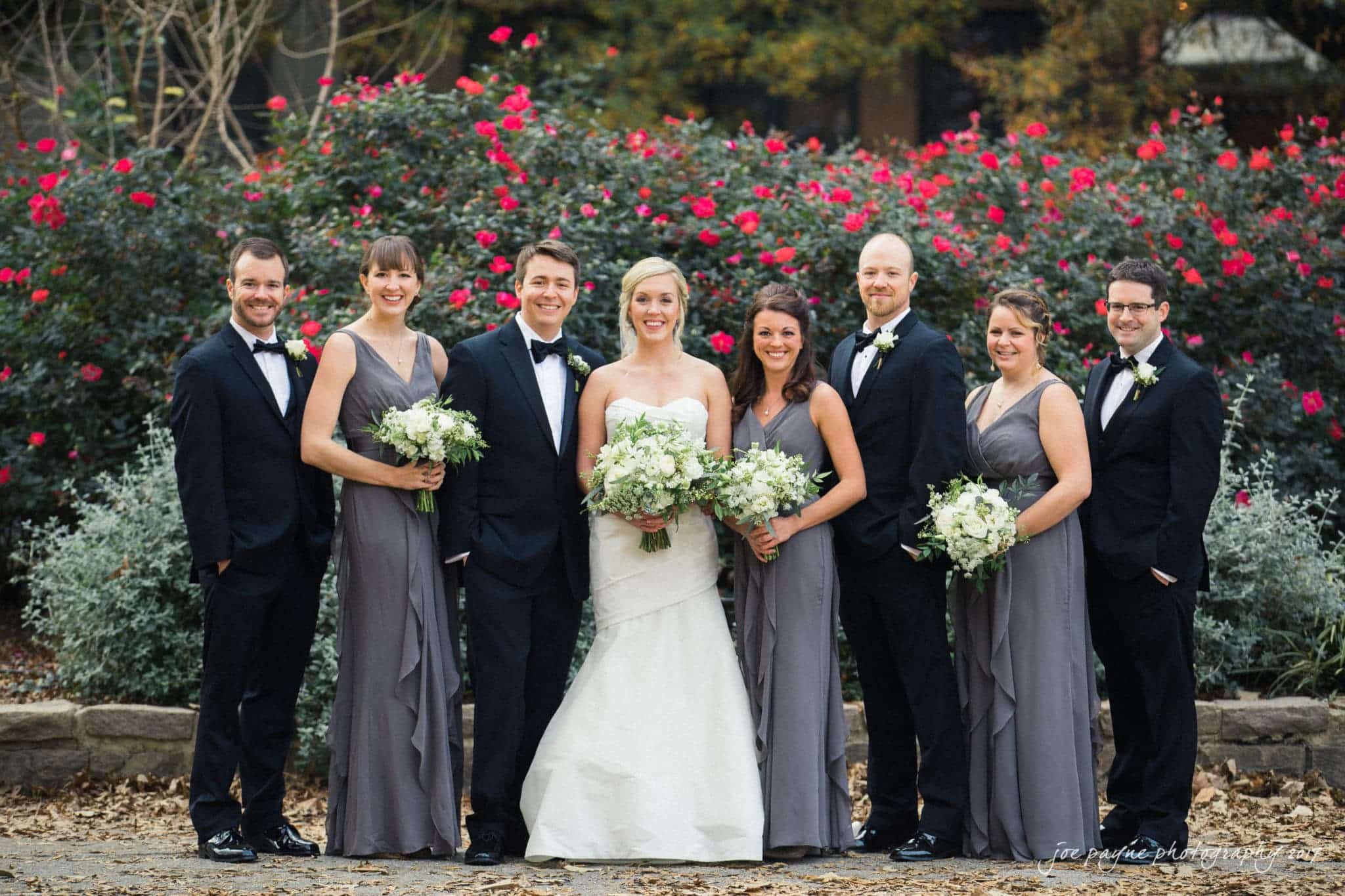 A bride and groom stand smiling with their bridal party in front of lush greenery and red flowers. The bride wears a white gown, and the bridesmaids wear gray dresses. The groom and groomsmen are in black tuxedos.
