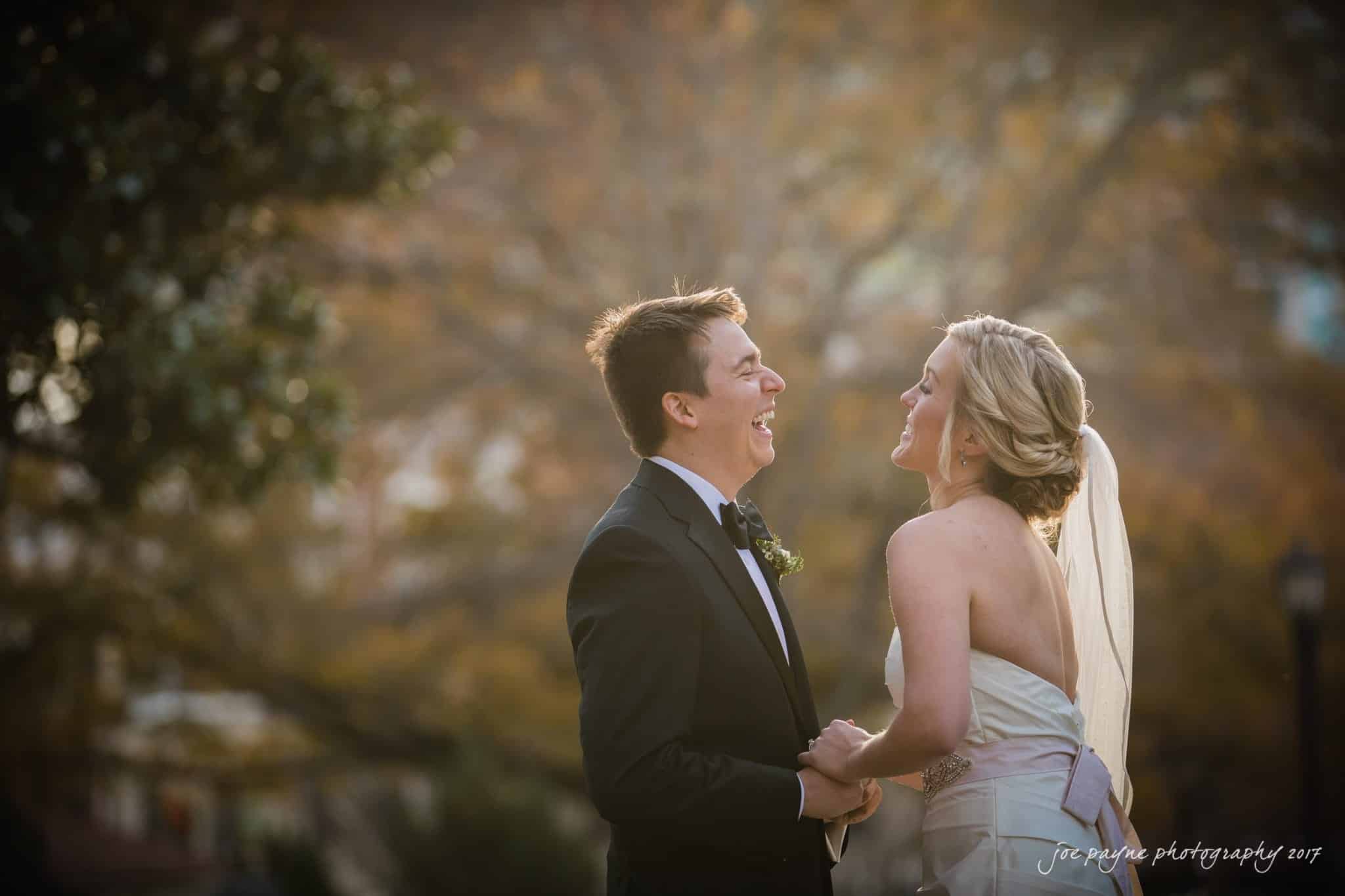 A bride and groom in formal attire share a joyful moment outdoors. The bride, in a white dress and veil, laughs while holding the grooms hands. The groom is in a black tuxedo, in front of a softly blurred autumn background.