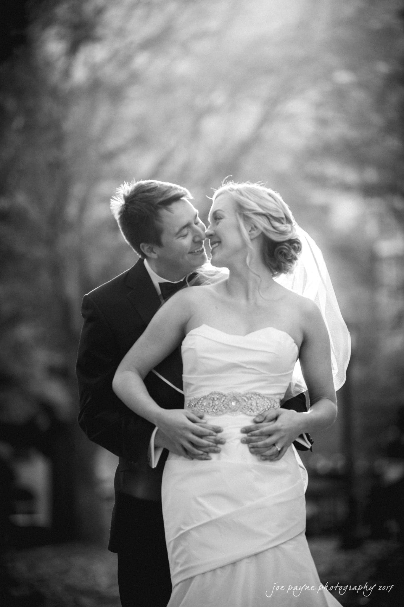 A black and white photo of a bride and groom smiling at each other. The bride is in a strapless dress with a veil, and the groom is in a suit. They are outdoors with blurred trees in the background, creating a warm, intimate atmosphere.