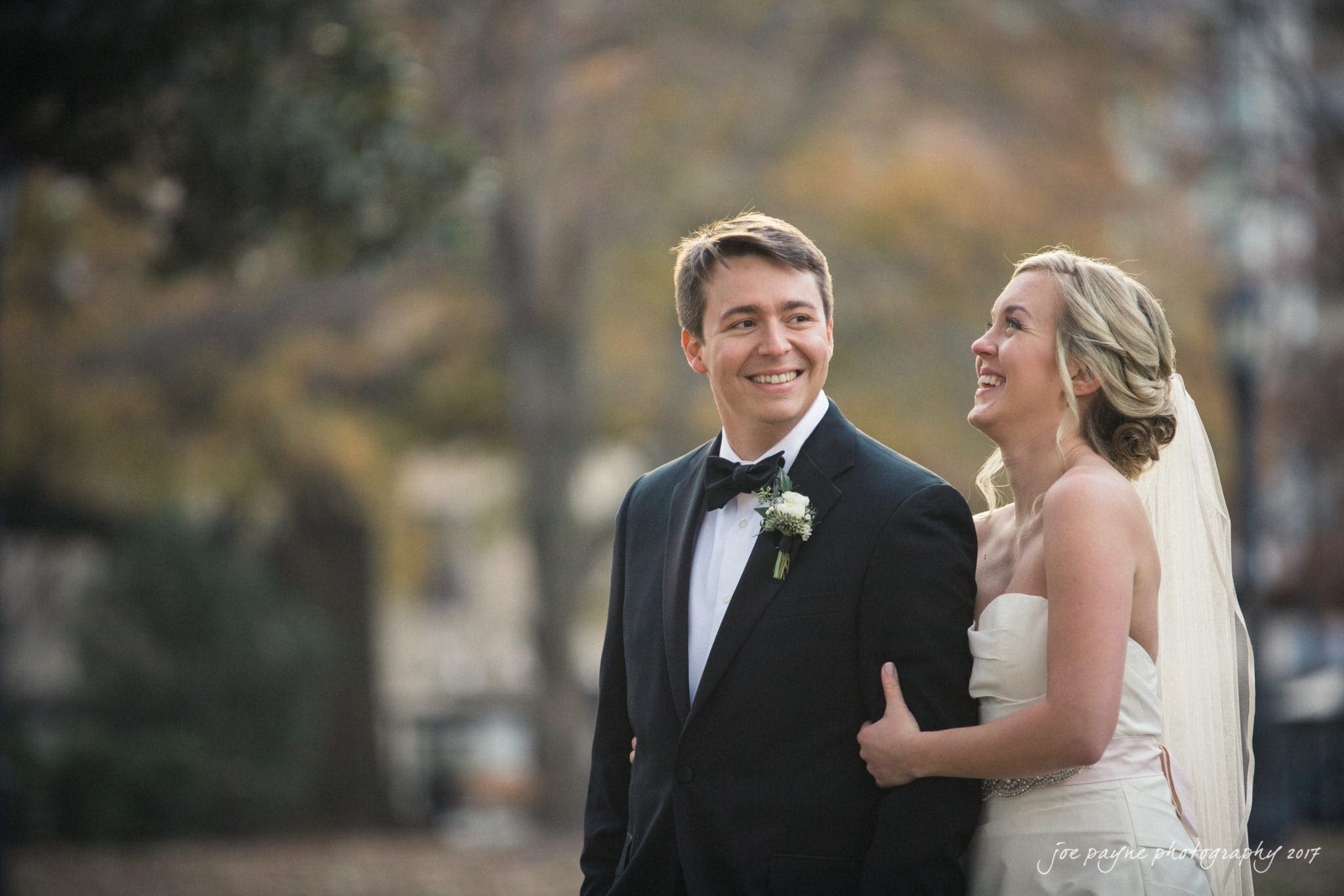 A bride and groom stand outside, smiling joyfully. The groom is in a black tuxedo with a bow tie, while the bride wears a strapless white gown and veil. They are embracing against a blurred background of greenery and soft autumn colors.