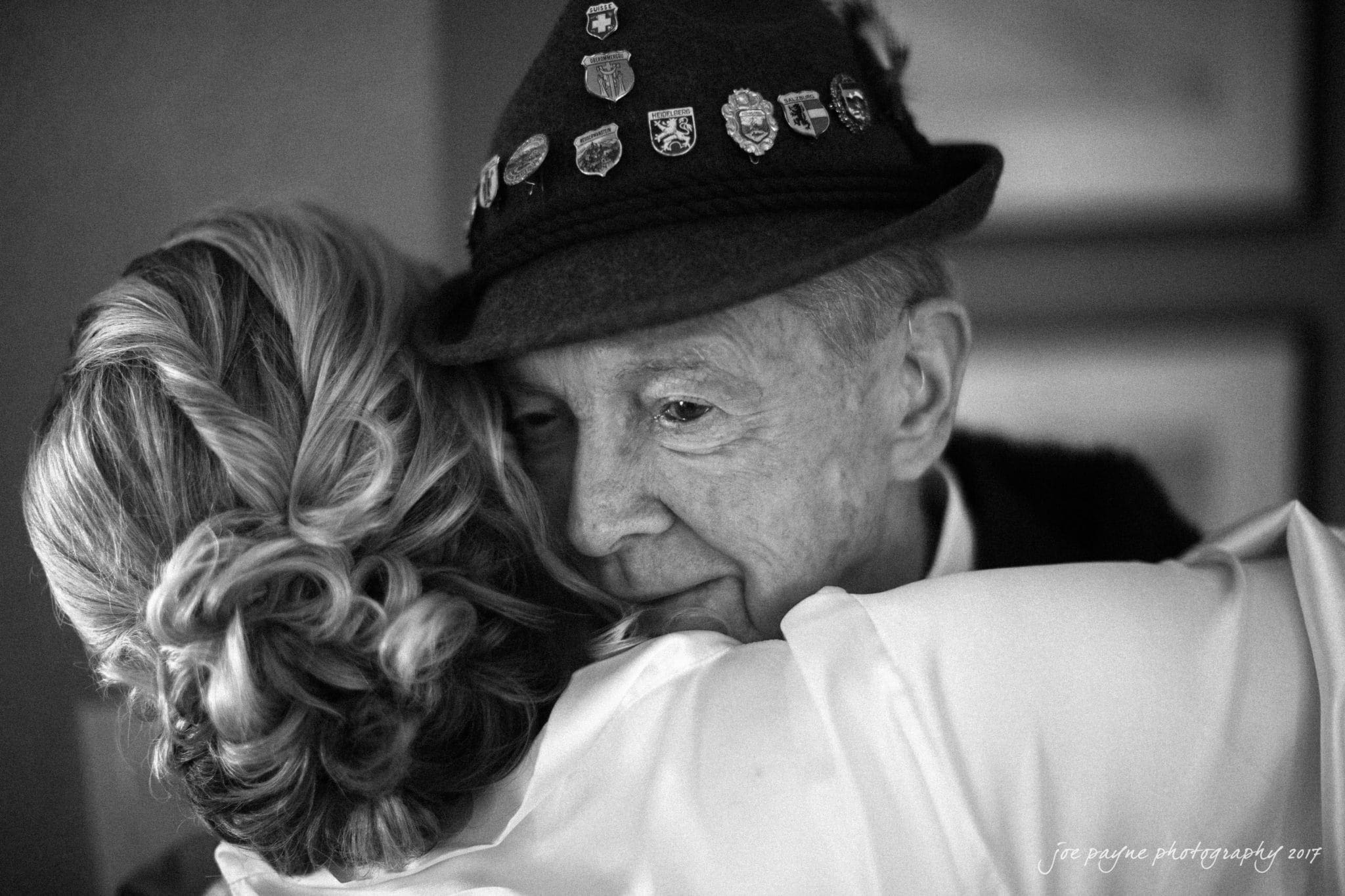 Black and white photo of a man in a hat decorated with pins hugging a woman. The man looks content and the woman has her back to the camera, showing her styled hair.